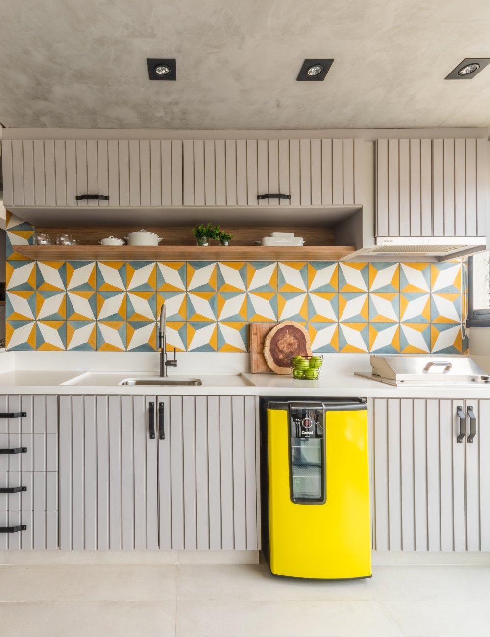 Cozinha moderna com armários cinza de ripas verticais e geladeira amarela retrô. O destaque fica por conta dos azulejos coloridos no backsplash, que adicionam um toque vibrante e contemporâneo ao ambiente. Estilo clean e funcional.