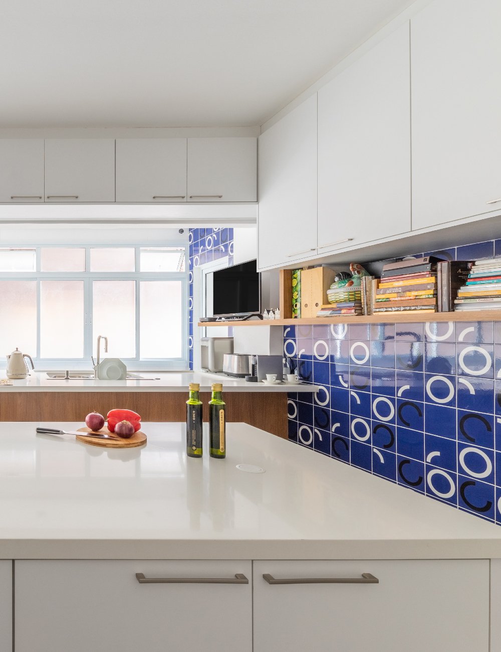 Cozinha moderna com armários brancos, bancada de mármore e detalhes em madeira clara. Azulejos coloridos com padrões geométricos dão um toque retrô. Itens decorativos e livros na prateleira. Detalhes minimalistas e clean.