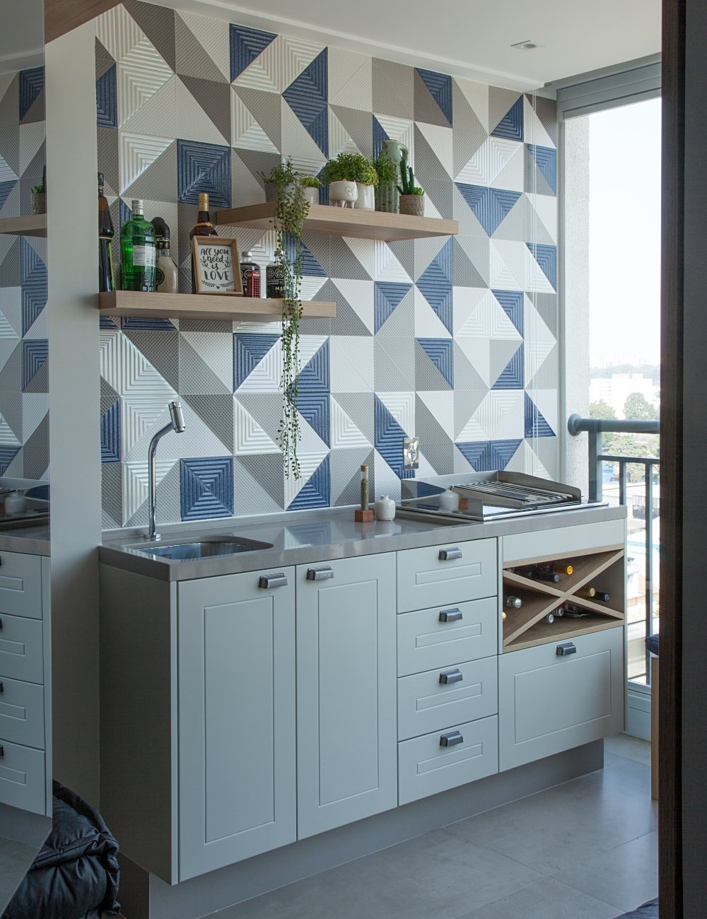 Cozinha moderna com armários brancos, bancada cinza e azulejos coloridos em tons de azul, cinza e branco. Prateleiras de madeira clara com plantas e bebidas. Tendência minimalista e cores neutras com toques de cor para sofisticação e modernidade.