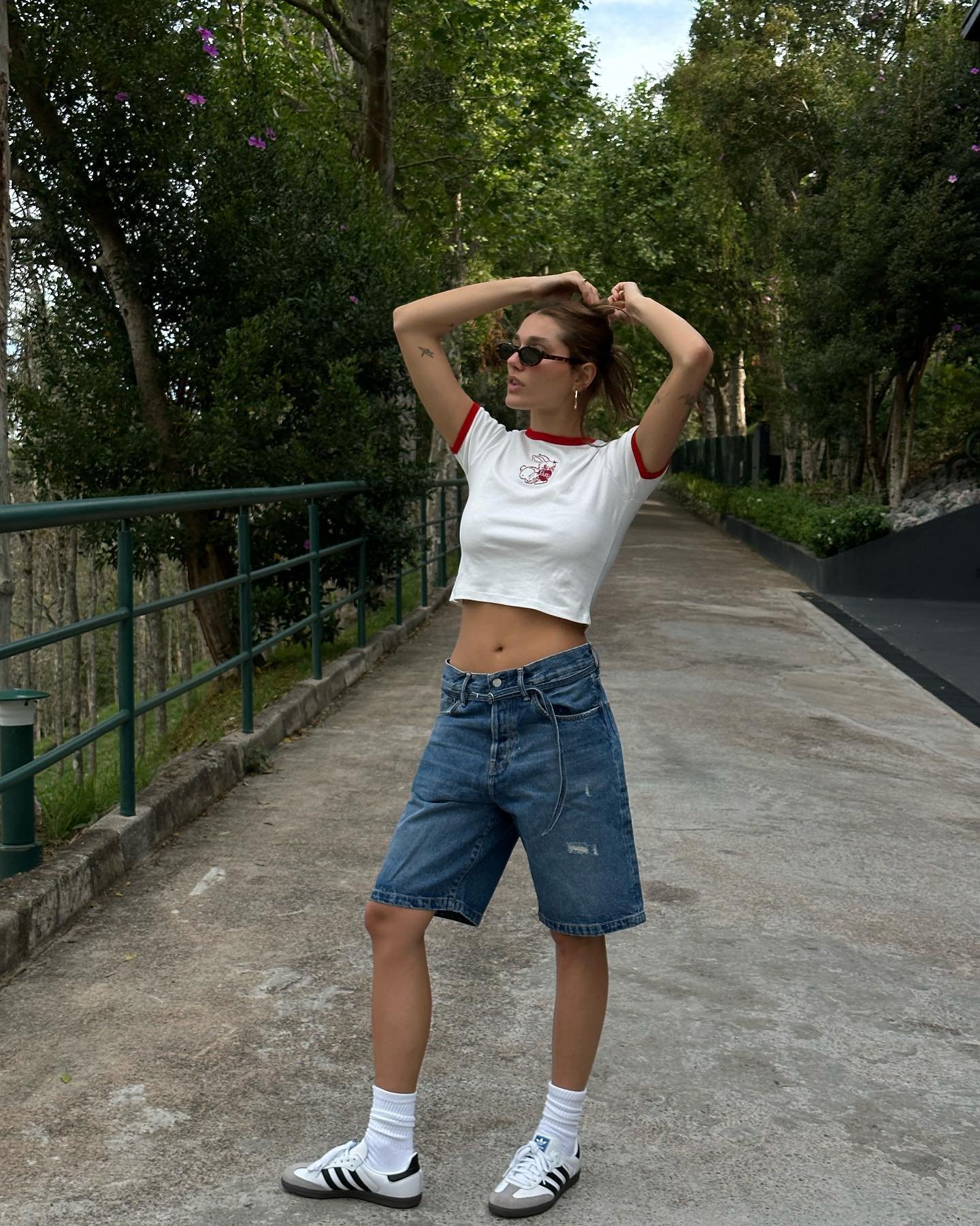 Jovem usa look casual com camiseta cropped branca de mangas curtas e detalhes em vermelho. Bermuda jeans azul de modelo largo. Nos pés, tênis branco com listras pretas, combinado com meias altas brancas.