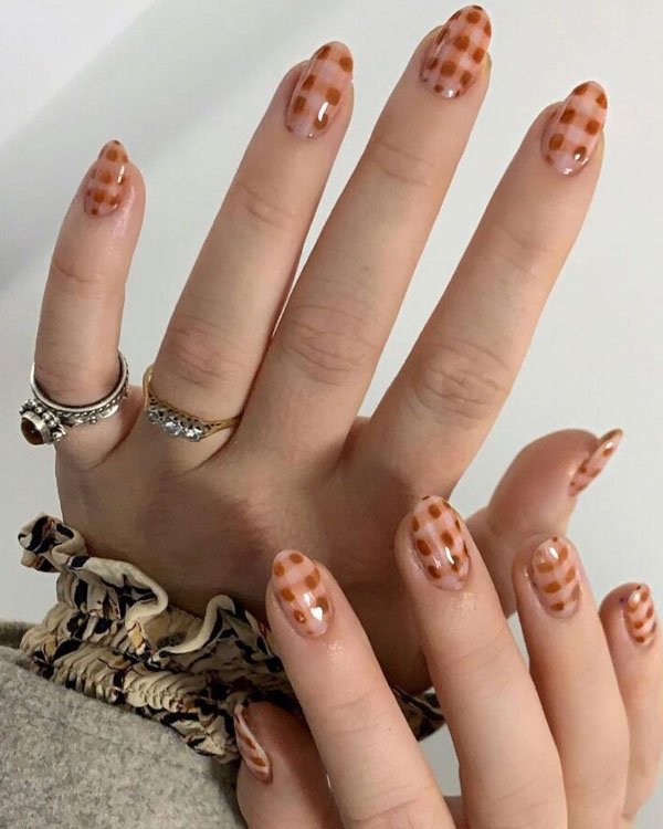 Unhas com esmalte xadrez marrom, tendência de nail art moderna. Mãos com anéis dourados e prateados, unhas ovais, combinação de estilo casual e elegante. Mangas de blusa com estampa sutil, contrastando com a nail art.