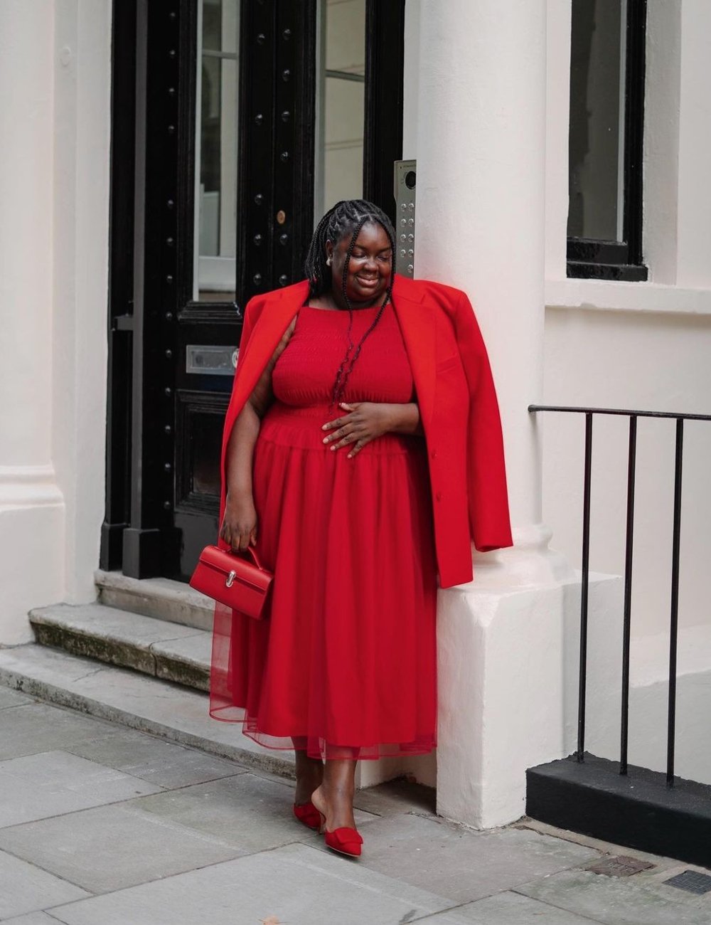 Mulher usando vestido midi vermelho, blazer oversized por cima dos ombros e sapatos de salto baixo vermelhos. Segura uma bolsa vermelha combinando. O conjunto monocromático destaca tendências de moda minimalista e elegante.