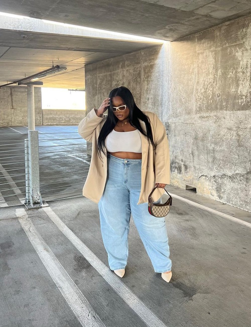 Mulher usando blazer oversized bege, cropped branco e calça jeans larga, acompanhados de óculos de sol brancos e bolsa pequena Gucci. Moda casual e confortável com toques de luxo e estilo urbano, destacando tendências oversized e acessórios de grife.