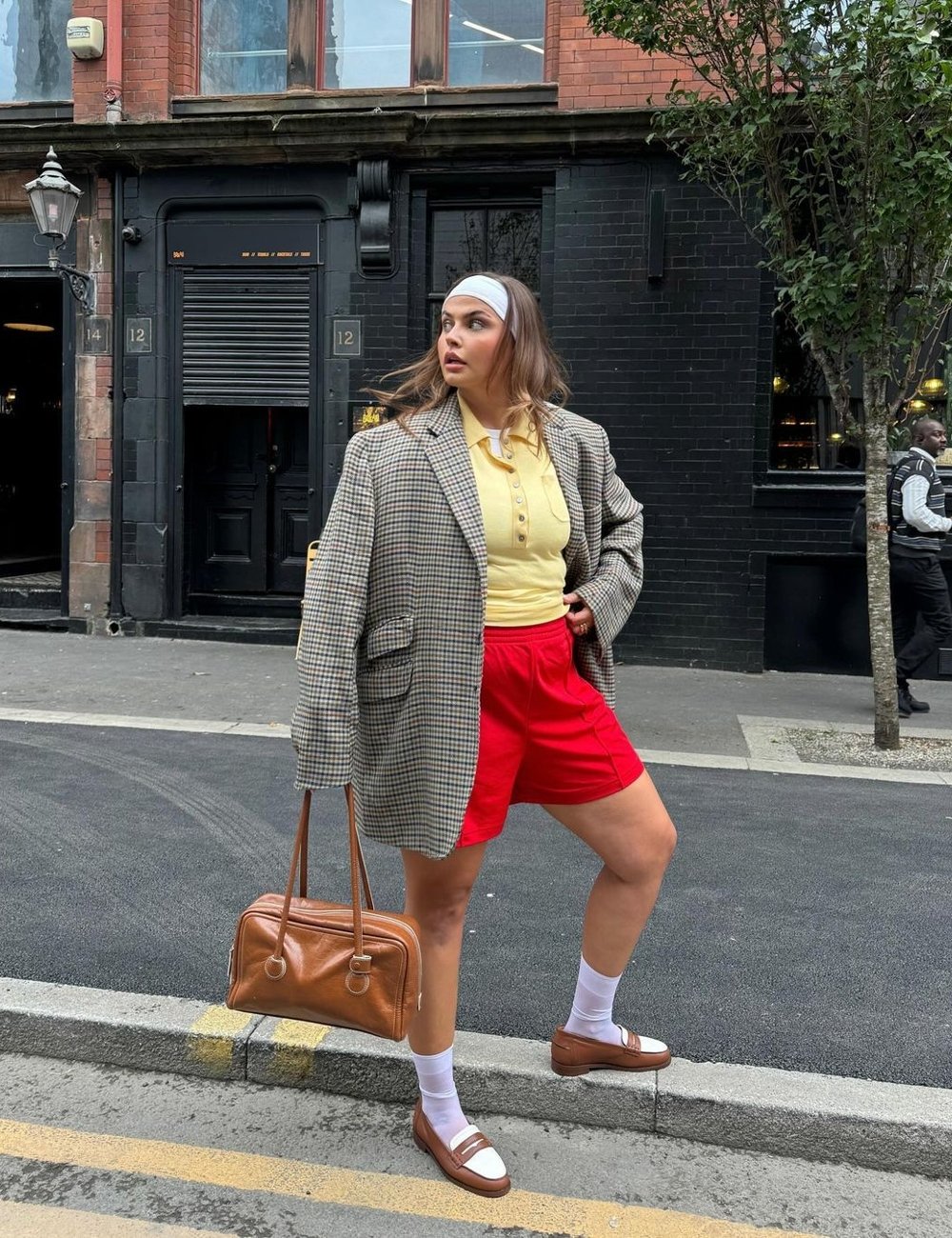 Mulher usando uma combinação eclética de tendências de moda: blazer oversized, polo amarela, shorts vermelhos, meias brancas e mocassins marrons. Completa o visual com uma faixa branca na cabeça e uma bolsa de couro marrom. Estilo casual e despojado.