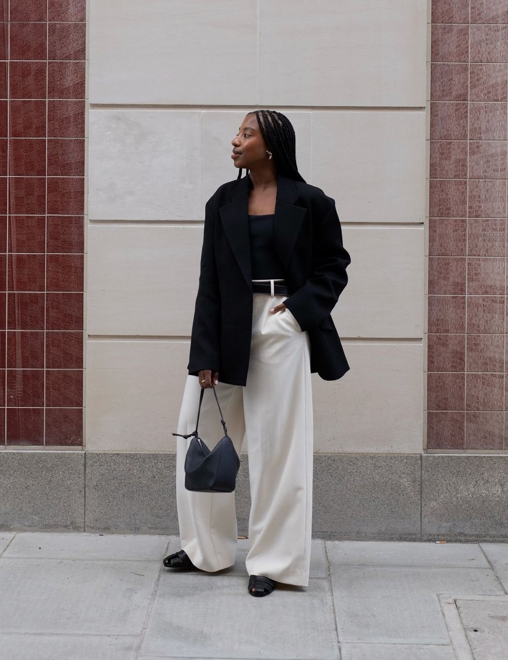 Look sofisticado e elegante: blazer oversized combinado com top preto, calça de cintura alta branca e sapatos estilo mocassim pretos. Acessórios incluem bolsa pequena preta e cabelos trançados, destacando tendências de moda minimalista e chique.