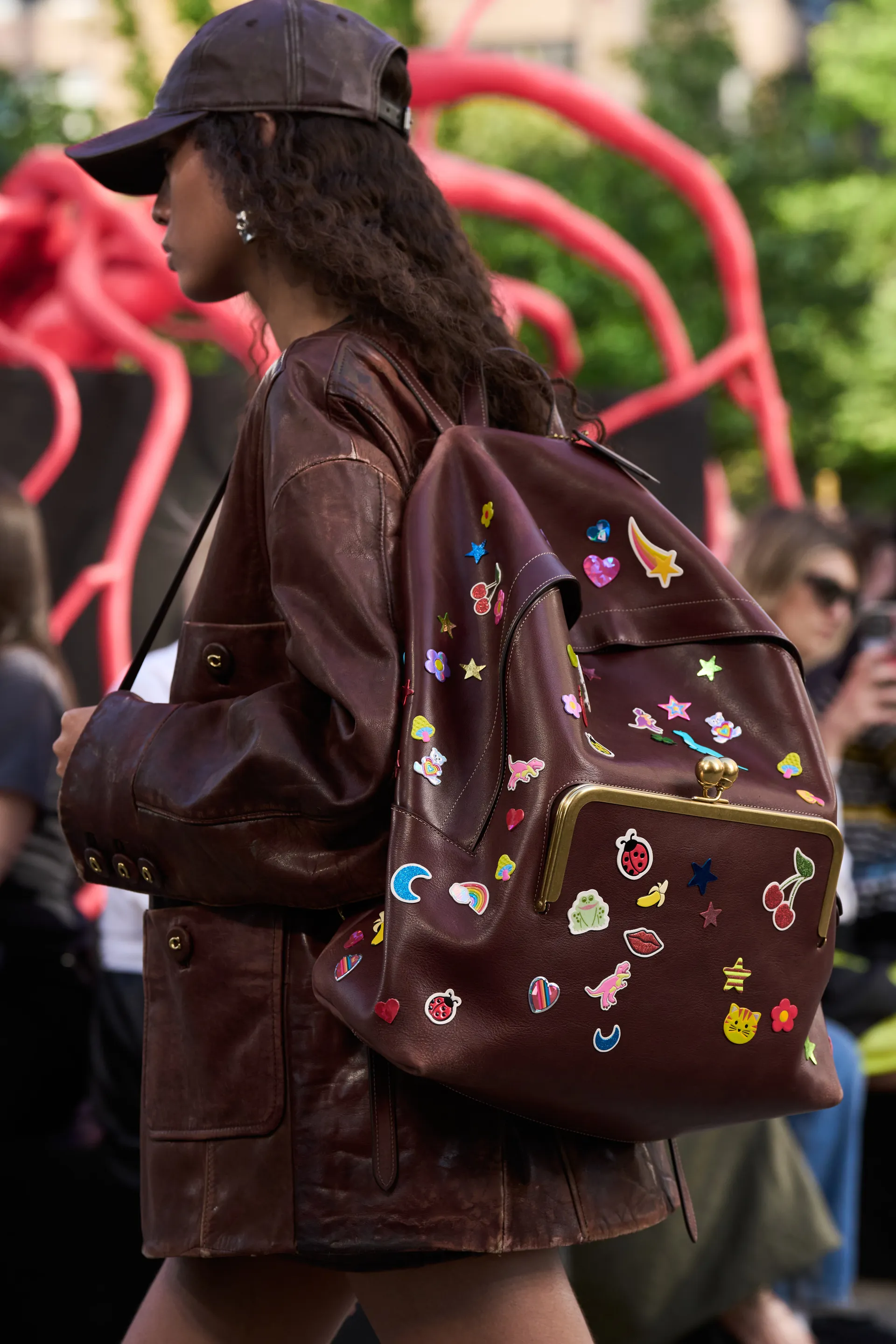 Jovem vestindo jaqueta de couro marrom oversized e boné combinando. Mochila de couro marrom decorada com adesivos coloridos, estilo lúdico e divertido. NYFW SS25