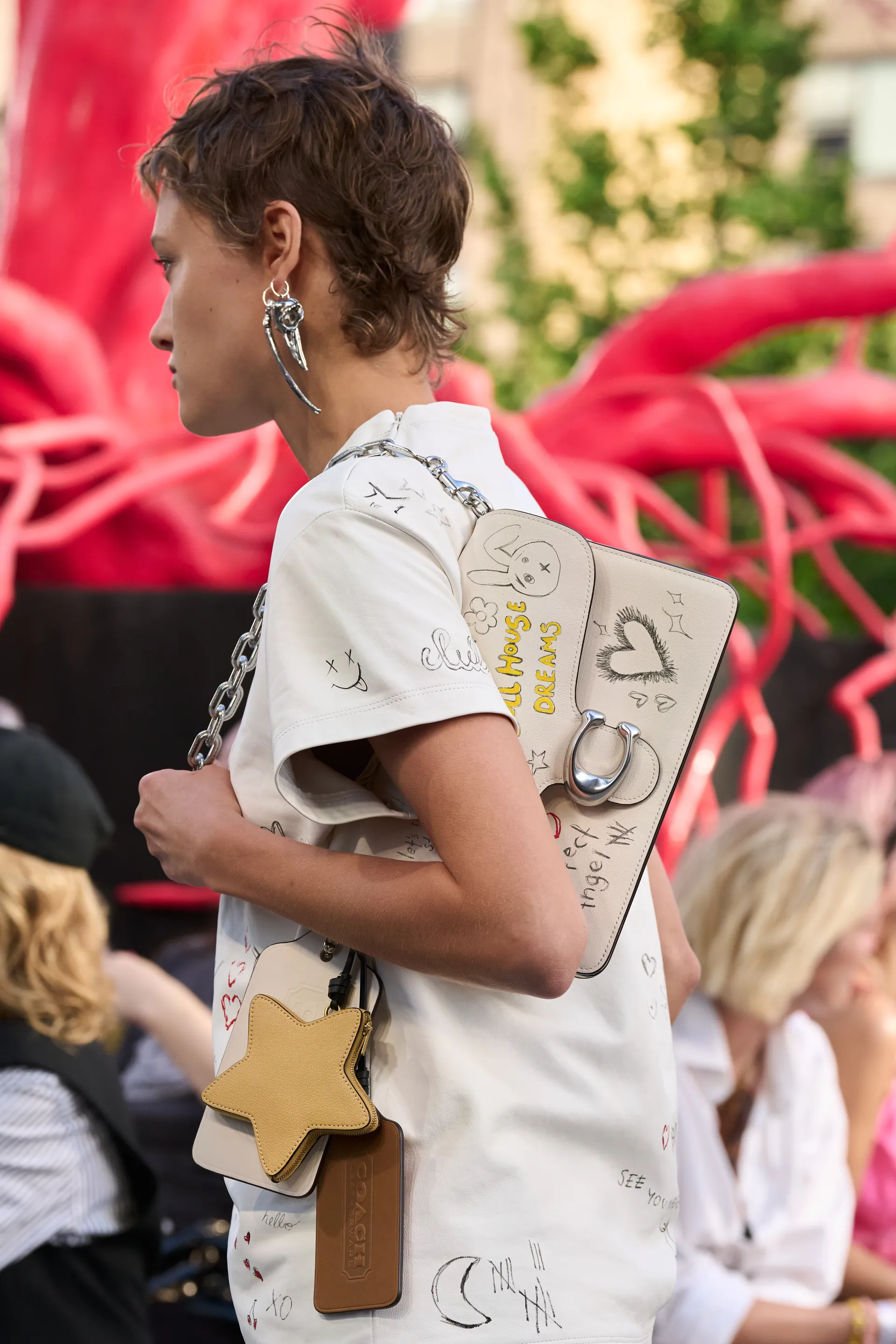 Pessoa com estilo moderno usando camiseta branca com desenhos e bolsa decorada com patches e alça de corrente. O visual é complementado por um brinco prateado grande e estiloso. Tendências de moda: acessórios statement e peças personalizadas.