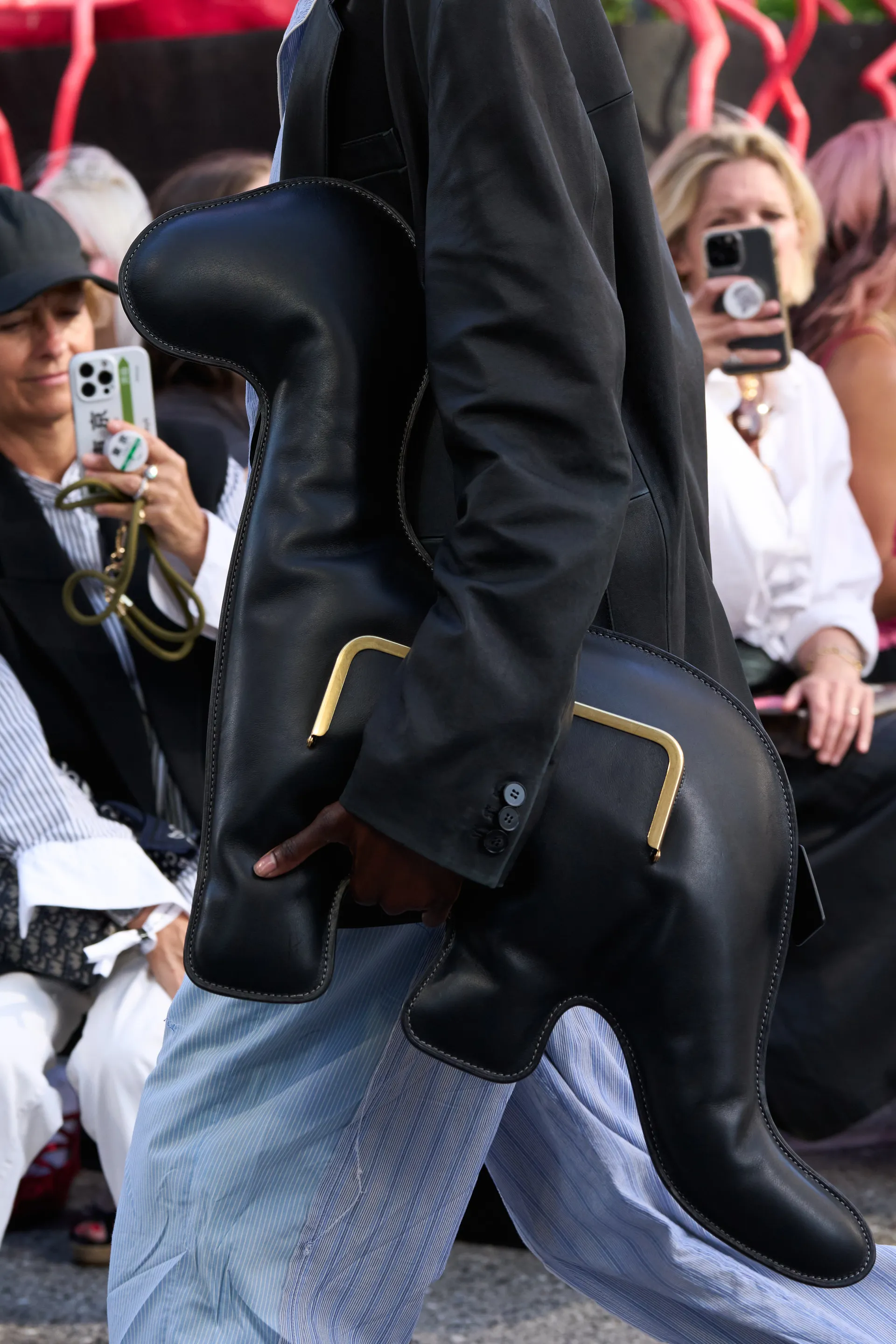 Desfile de moda com destaque para bolsa preta em formato único e estrutura inovadora. Traje composto por blazer preto de alfaiataria e calça listrada azul clara, combinando elegância e tendências contemporâneas. Fotógrafos e público em segundo plano.