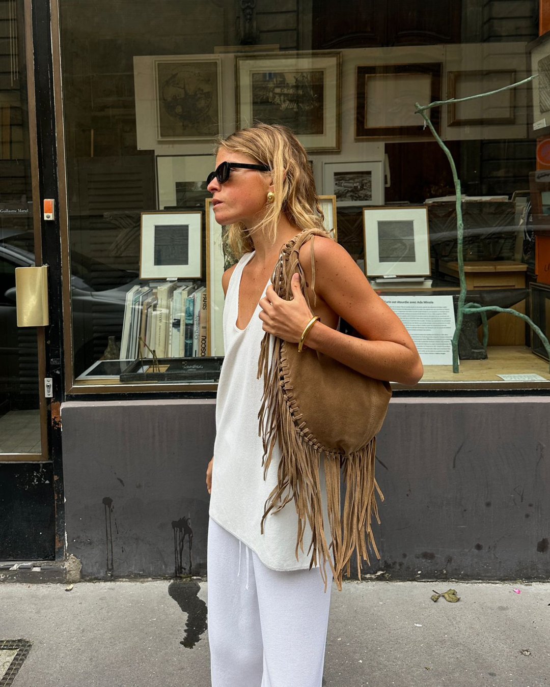 Mulher vestida com look casual e estiloso, usando regata branca e calça larga também branca. Acessórios incluem óculos de sol preto, bolsa de franja e brincos dourados. Tendências: minimalismo e boho chic.