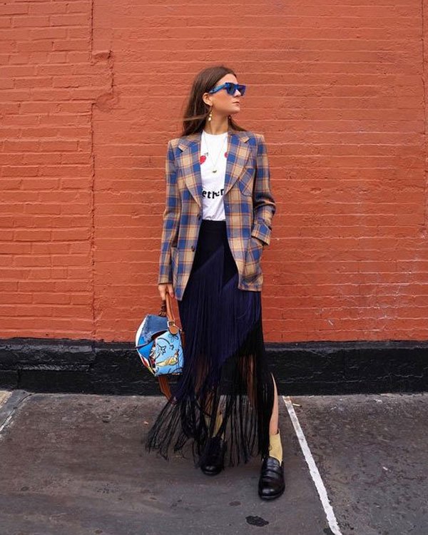 Mulher com look fashionista, combinando blazer de lã xadrez, camiseta branca casual e saia preta com franjas. Acessórios incluem óculos de sol azul e bolsa estampada divertida.