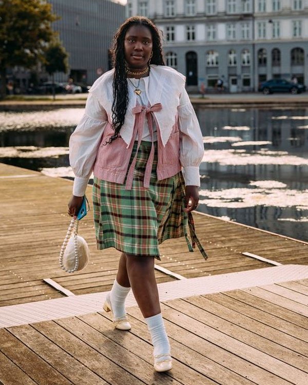 Mulher com look eclético: blusa branca bufante, colete rosa de babados, saia xadrez verde, meia-calça branca 3/4 e sapatilhas creme. Acessórios incluem colar dourado e bolsa de pérolas. Combinação de tendências vintage e modernas.