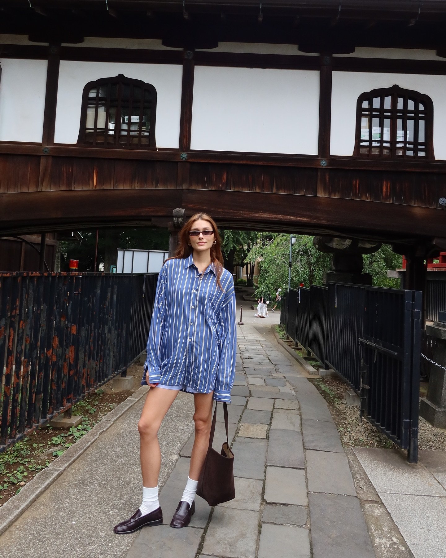 Jovem vestida com camisa oversized listrada azul e branca, shorts curtos, bolsa de ombro preta, meias brancas e mocassins marrom escuro. Estilo casual-chic, tendência de moda urbana e despojada. Óculos de sol completam o look contemporâneo.