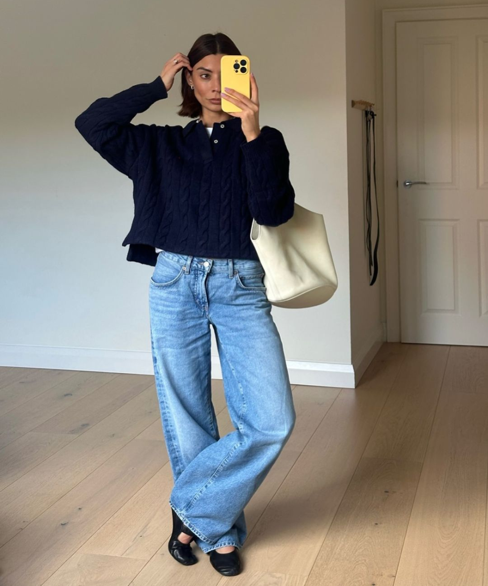 Mulher vestindo suéter azul marinho, calça jeans de perna larga de cintura alta e sapatilha preta. Acessório: bolsa branca grande. Look casual e sofisticado, tendência oversized com mistura de peças clássicas. Crítica de moda de outono-inverno.