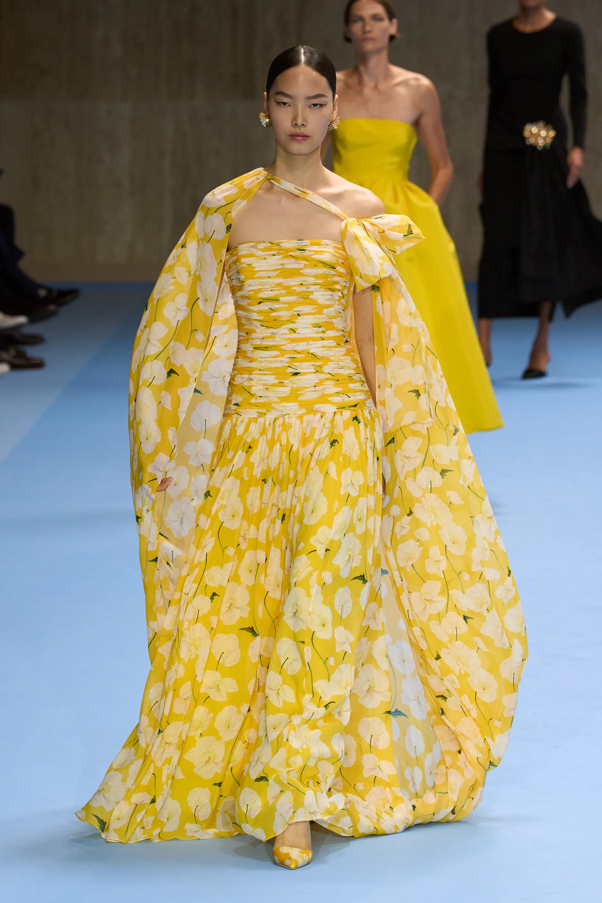 Vestido longo amarelo com estampa floral branca, destacando um ombro só e um laço chamativo no ombro esquerdo. Complementado por uma capa fluida no mesmo tecido. Look sofisticado e elegante, ideal para eventos formais e desfiles de moda.