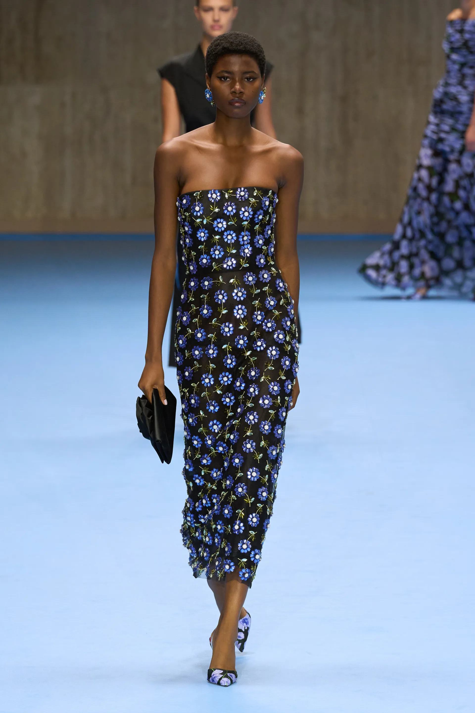 Modelo vestindo um elegante vestido midi tomara que caia decorado com flores azuis, exibindo uma tendência com transparências e bordados detalhados. Acompanhado de brincos combinando e uma clutch preta, destacando características da alta moda contemporânea.