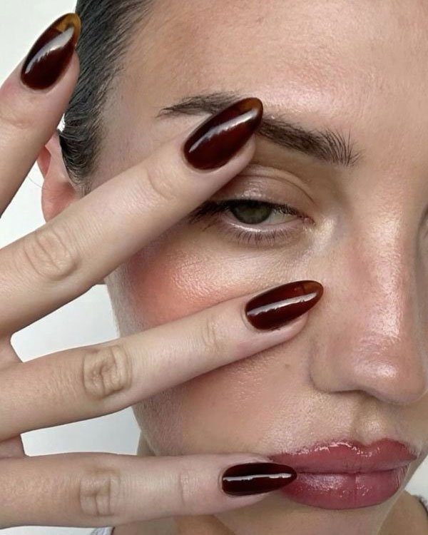 Foto em close-up de mulher destacando tendências de moda de unhas. As unhas estão longas, com formato amendoado, e pintadas em esmalte marrom brilhante.