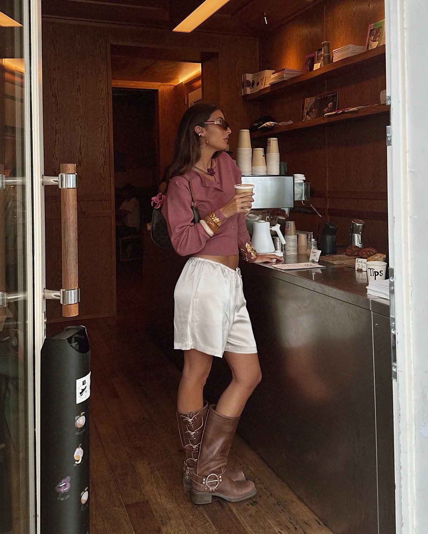 Mulher em cafeteria vestindo uma blusa roupa mauve de manga longa, short branco de cintura alta e botas marrons estilo Western. Acessórios incluem óculos de sol e pulseiras douradas. Estilo casual e despojado, com influências country urbanas.