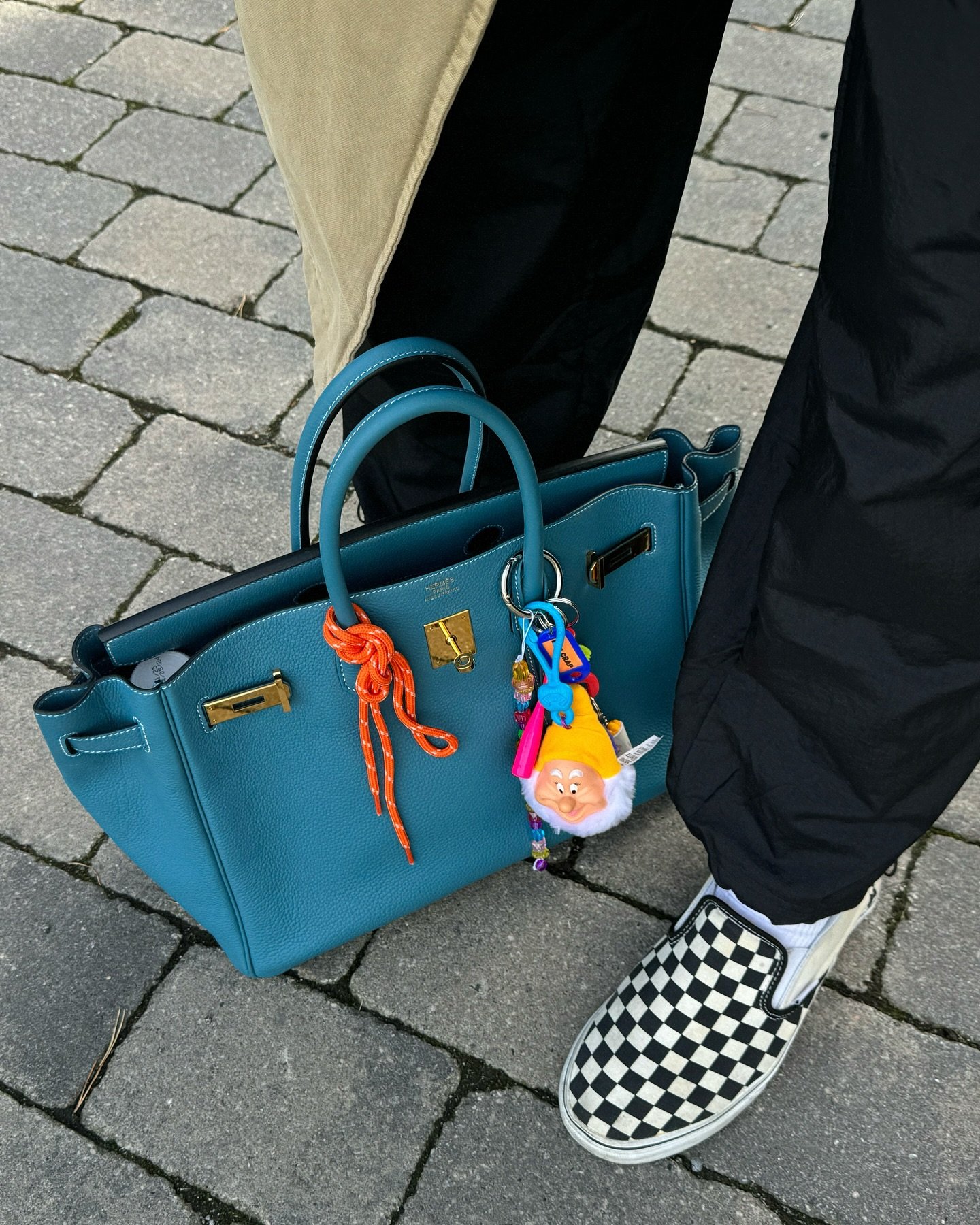 Bolsa de couro azul com amuleto colorido em um estilo despojado e personalizado. Tênis slip-on xadrez preto e branco complementam o look casual. Calça preta e casaco em tom neutro. Acessórios divertidos contrastam com peças clássicas.