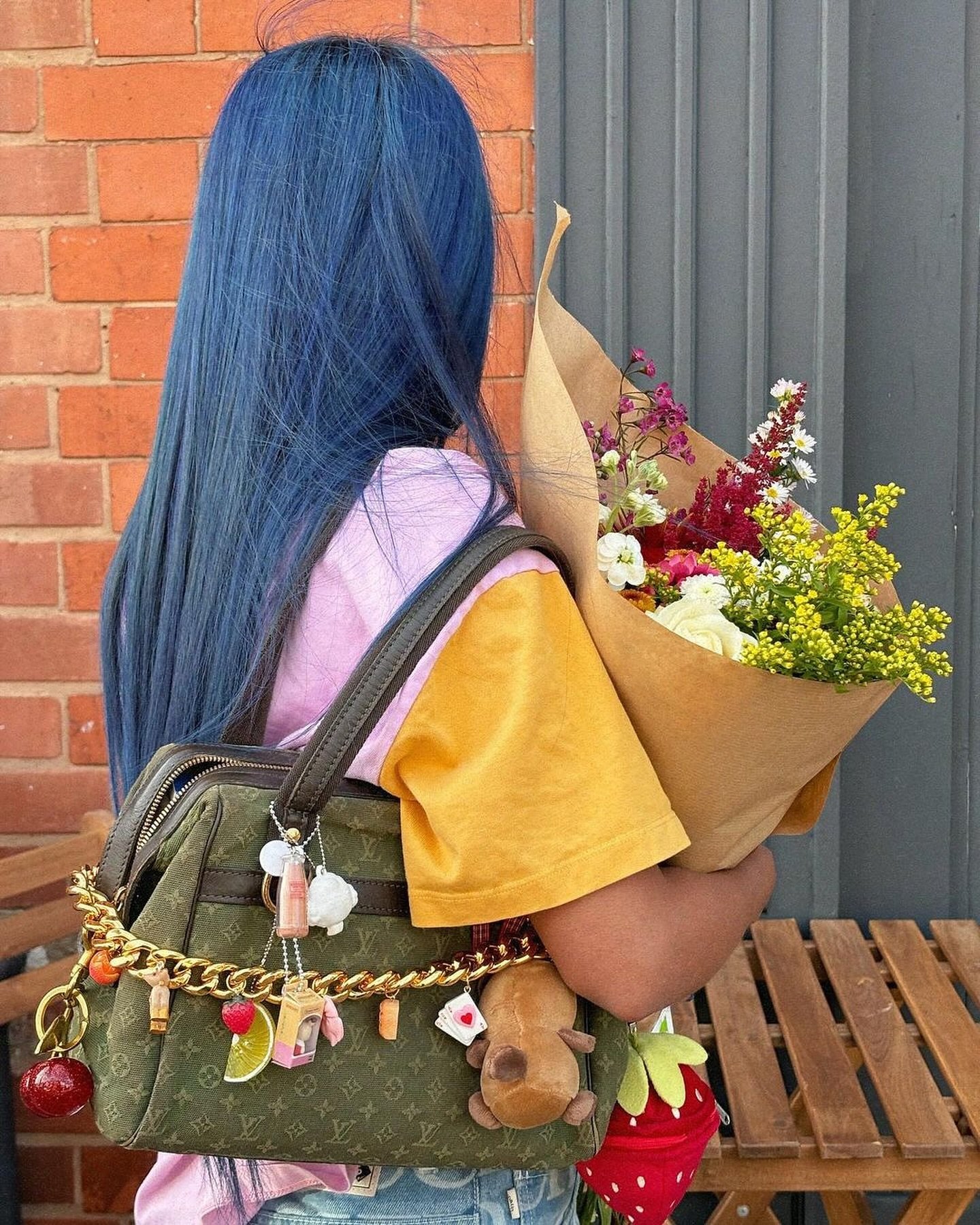 Pessoa com cabelo azul-vivo, vestindo camiseta bicolor rosa e amarela, segurando um buquê de flores. Bolsa de grife verde com chaveiros pendurados. Look casual e moderno com destaque para cabelo colorido e acessórios personalizados.