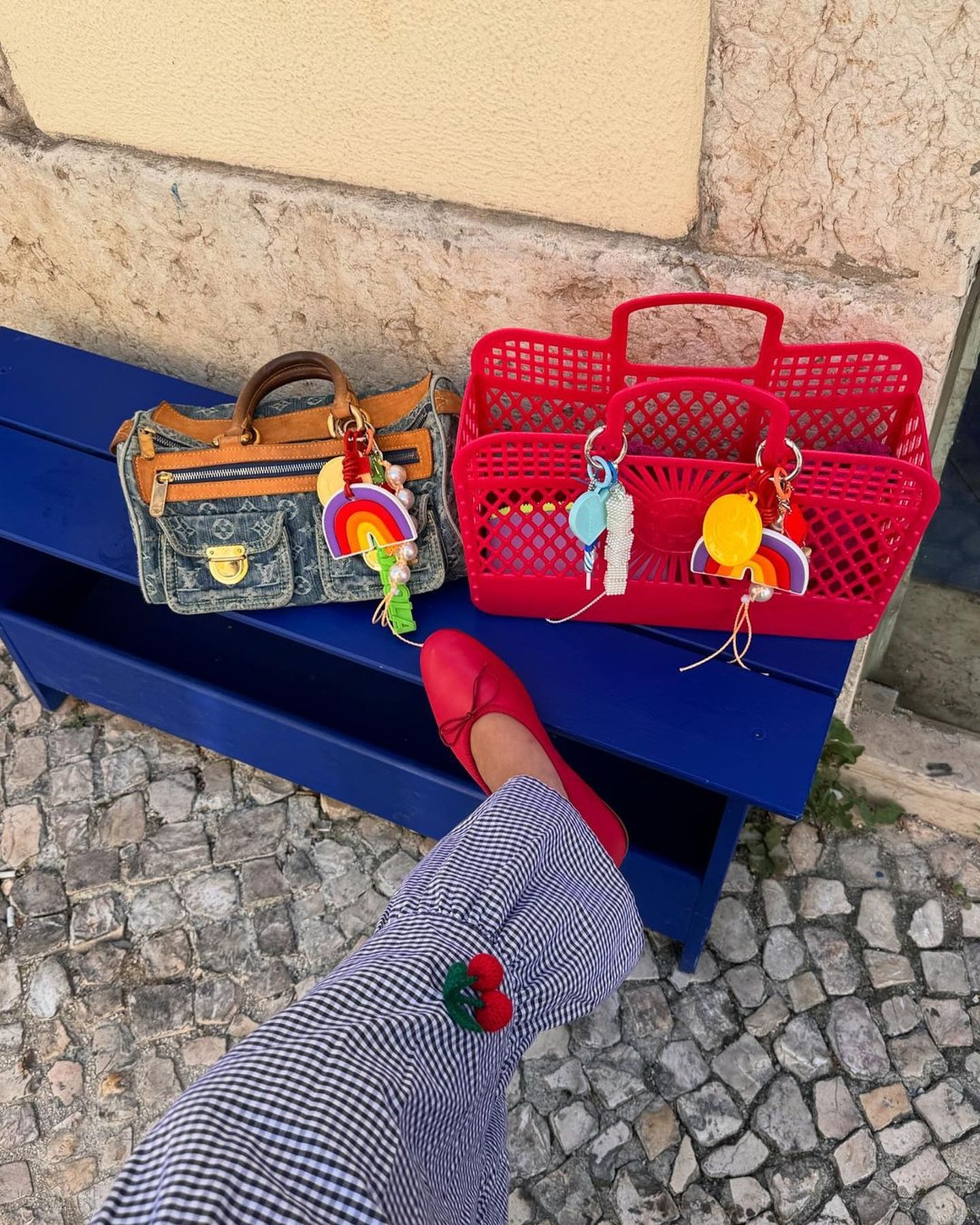 Foto de uma calça xadrez com bordado de cereja, combinada com sapatilha vermelha. Bolsas estilosas, incluindo uma jeans com detalhes em couro e outra plástica vermelha, adornadas com enfeites coloridos e chaveiros de arco-íris. Estilo casual e vibrante.