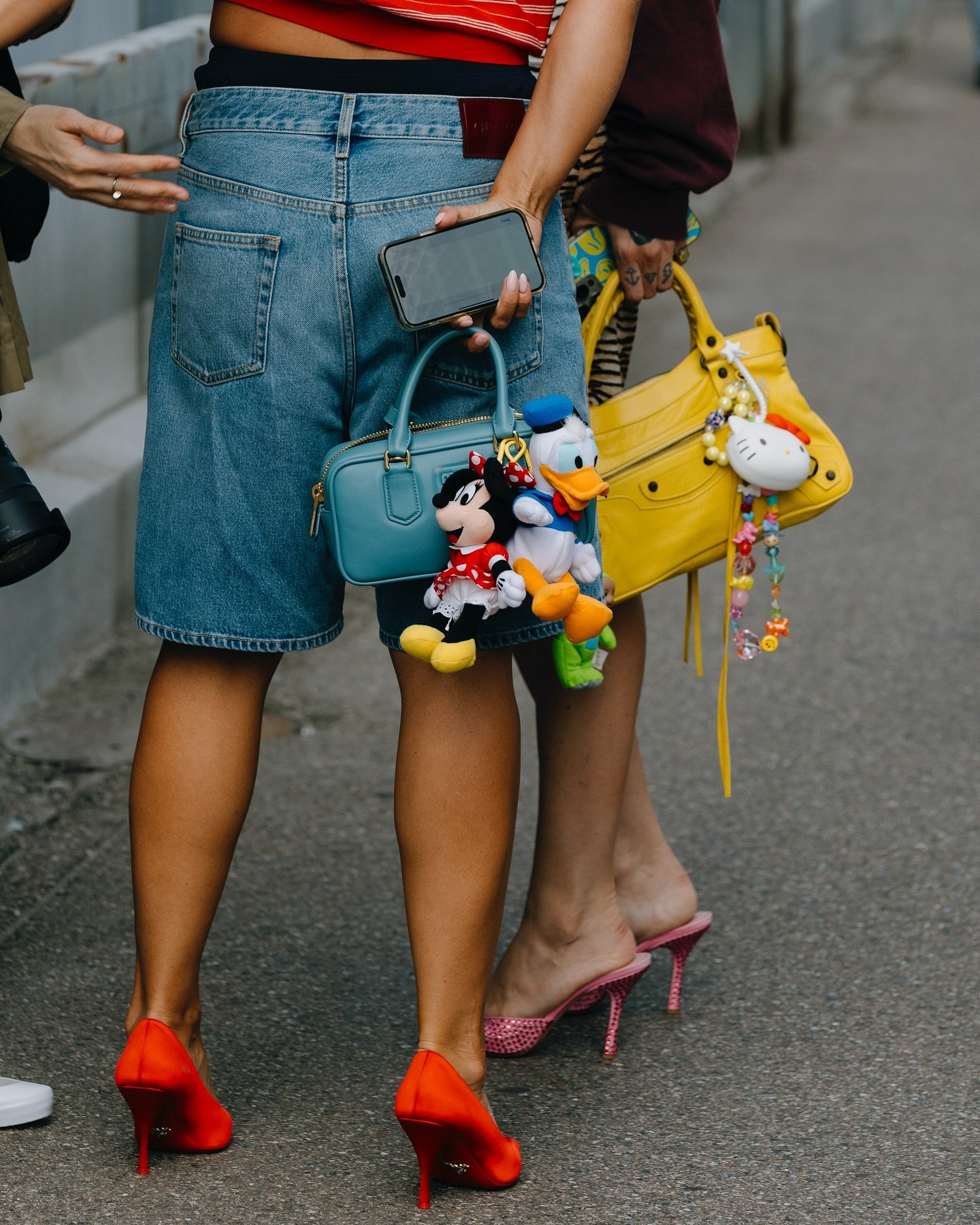 Moda urbana descontraída com shorts jeans largos e top cropped listrado. Acessórios incluem mini bolsas coloridas, chaveiros de personagens e sapatos de salto alto em tons vibrantes, como vermelho e rosa. Estilo jovem e divertido.