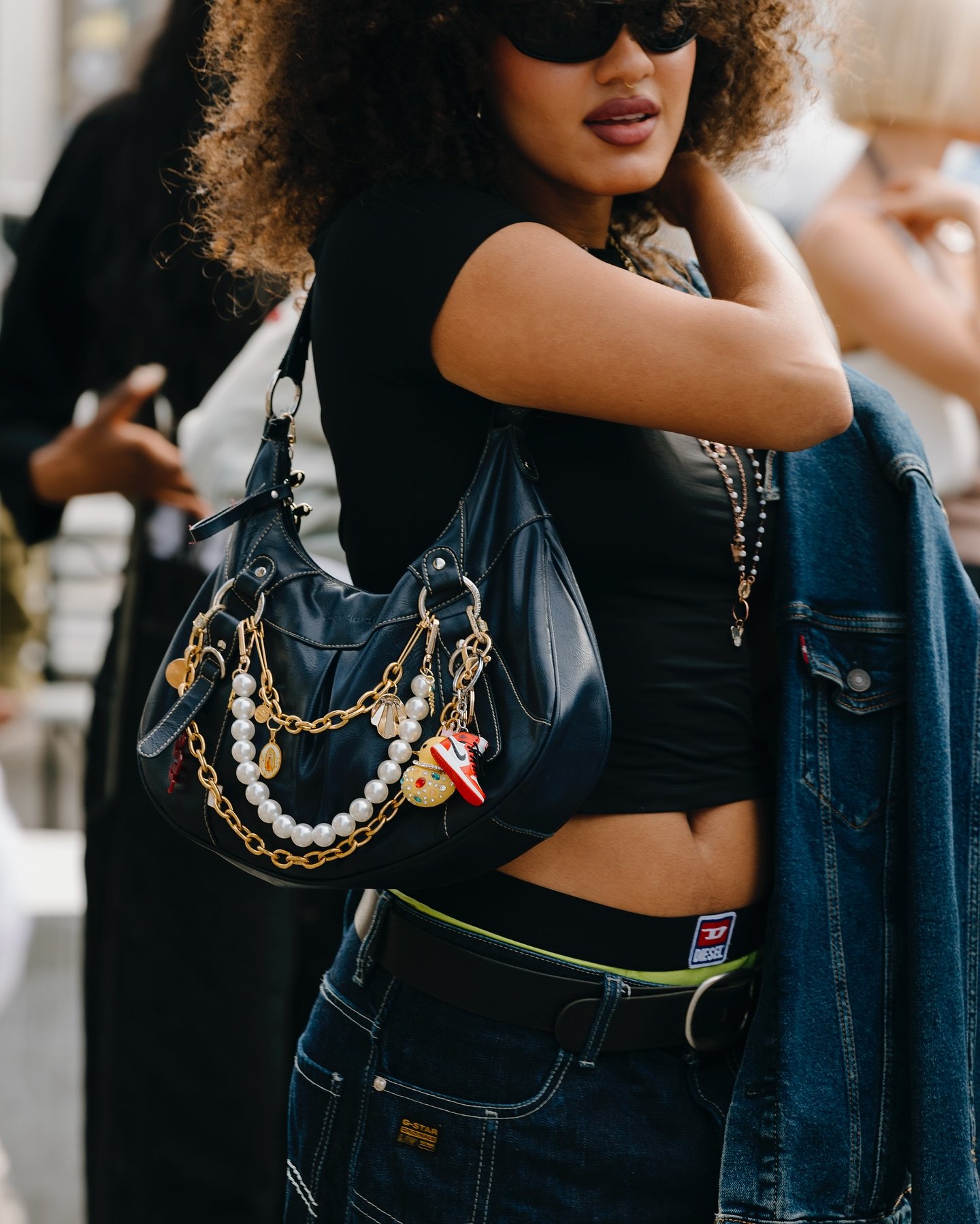 Mulher vestindo camiseta preta cropped e jeans de cintura baixa. Ela carrega uma bolsa preta decorada com correntes douradas e pérolas. Um casaco jeans está pendurado no braço, e ela usa óculos de sol. Estilo casual e moderno, com acessórios chamativos.