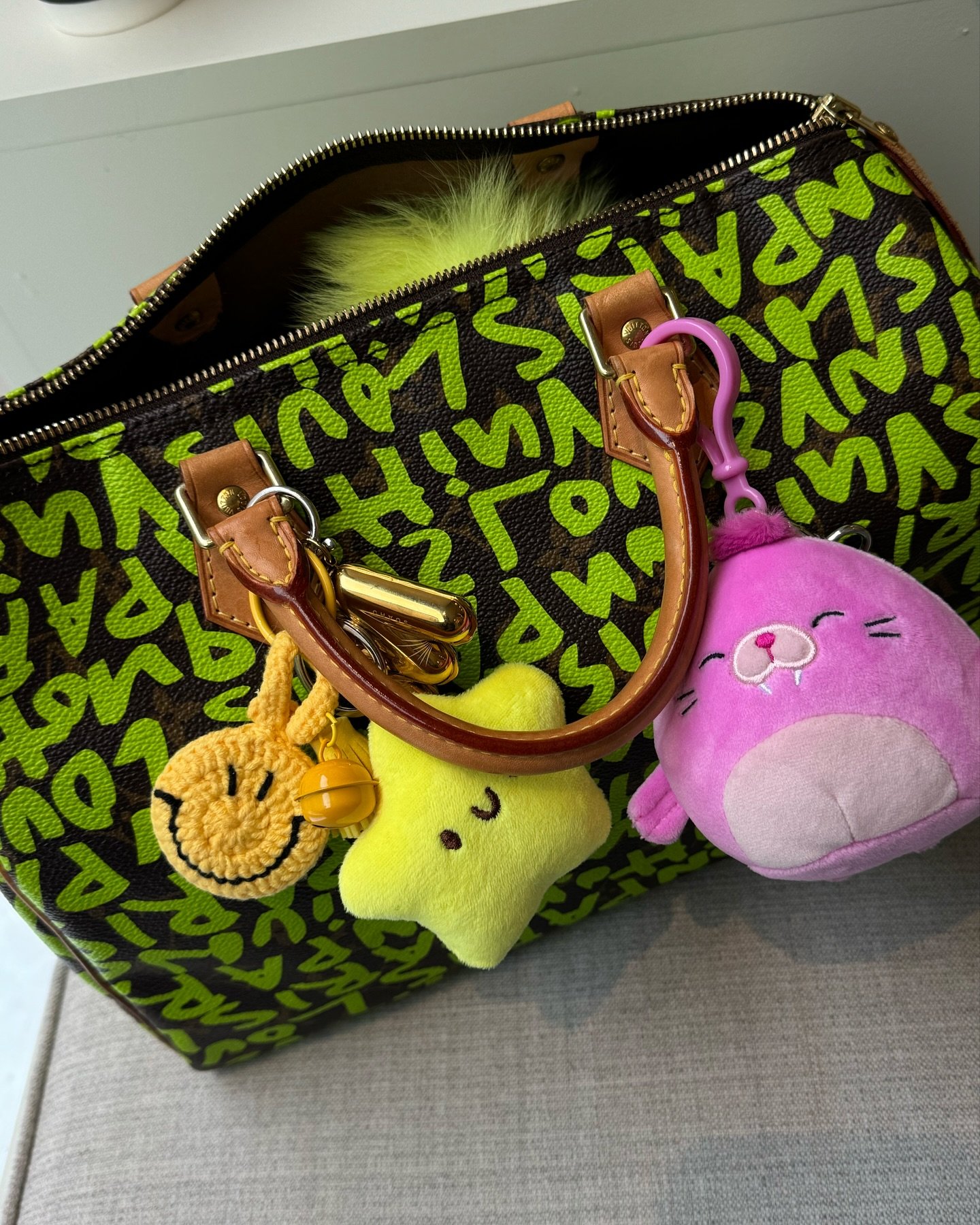 Bolsa estilosa com estampa gráfica verde e preta, alças de couro marrom e detalhes dourados. O acessório destaca-se pelos chaveiros de pelúcia divertidos: uma estrela amarela, um ursinho roxo e um emoji sorridente amarelo croche. Tendência jovem e descontraída.