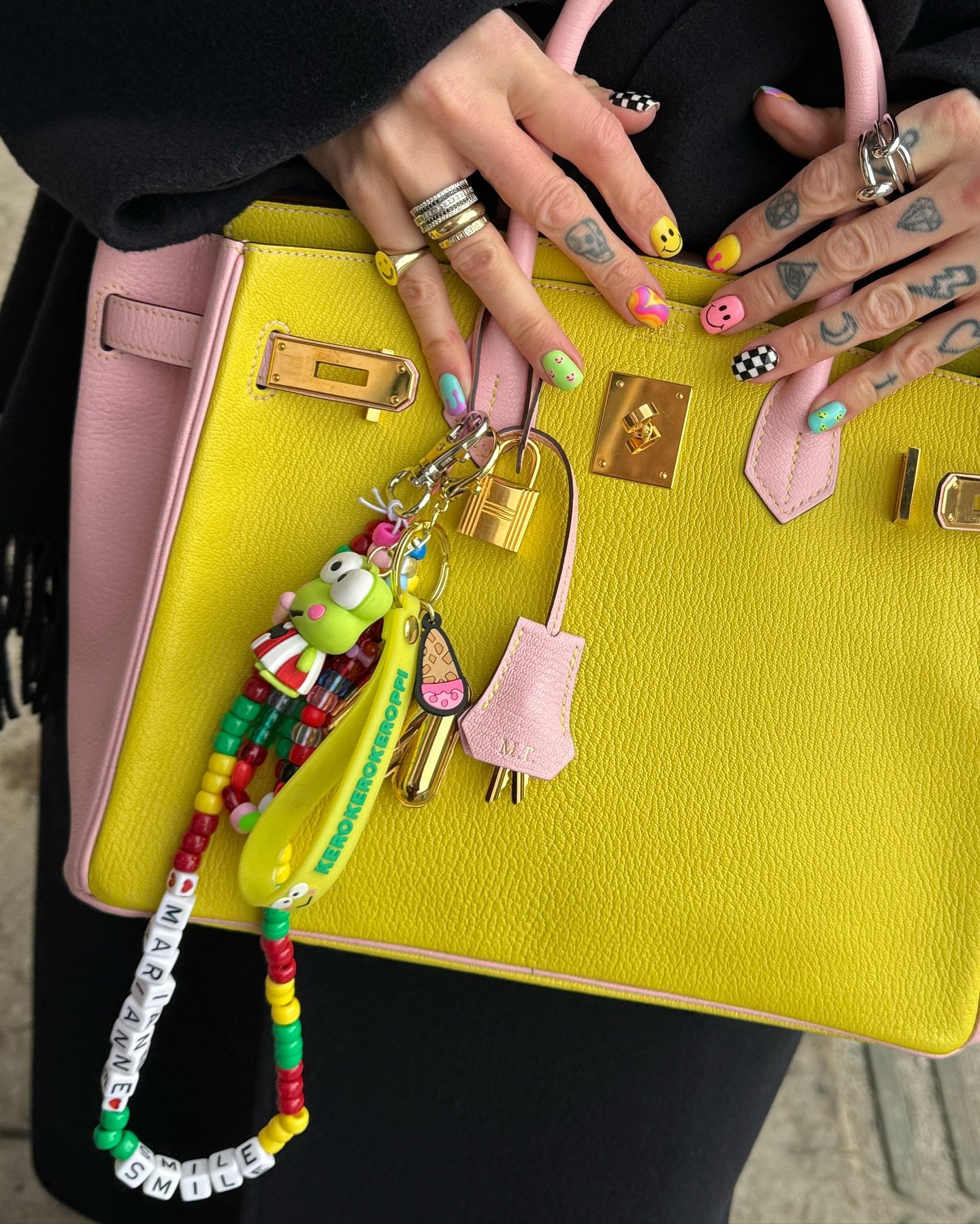 Mão com diversas tatuagens e unhas pintadas em estilo colorido e divertido, segurando uma bolsa de couro amarela com detalhes rosa. A bolsa possui charms e chaveiros com personagens animados, refletindo uma tendência de moda vibrante e personalizada.