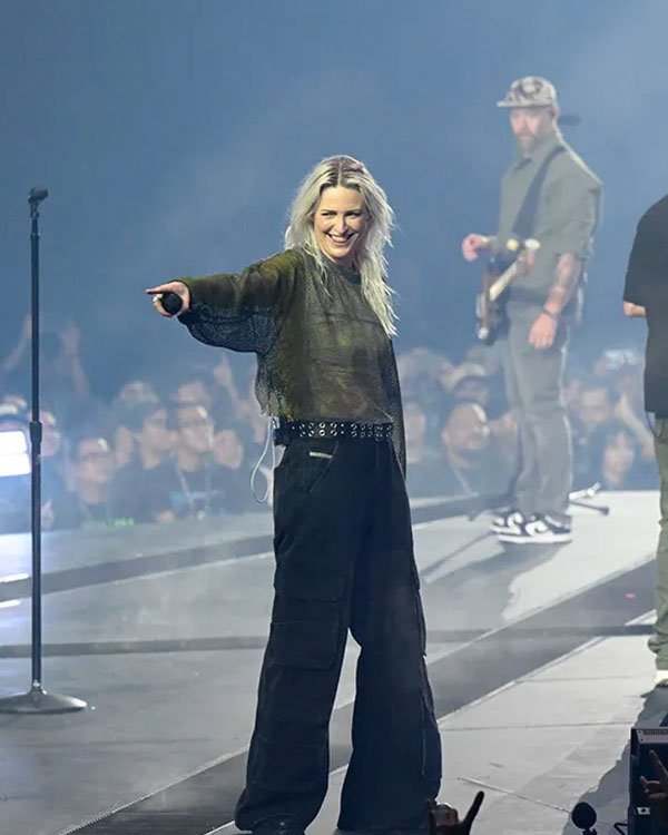 Imagem de artista no palco, vestindo look despojado: blusa transparente manga longa verde escura, combinada com calça cargo preta de cintura alta e cinto de tachinhas. Estilo urbano e contemporâneo, popular em shows e eventos de música.