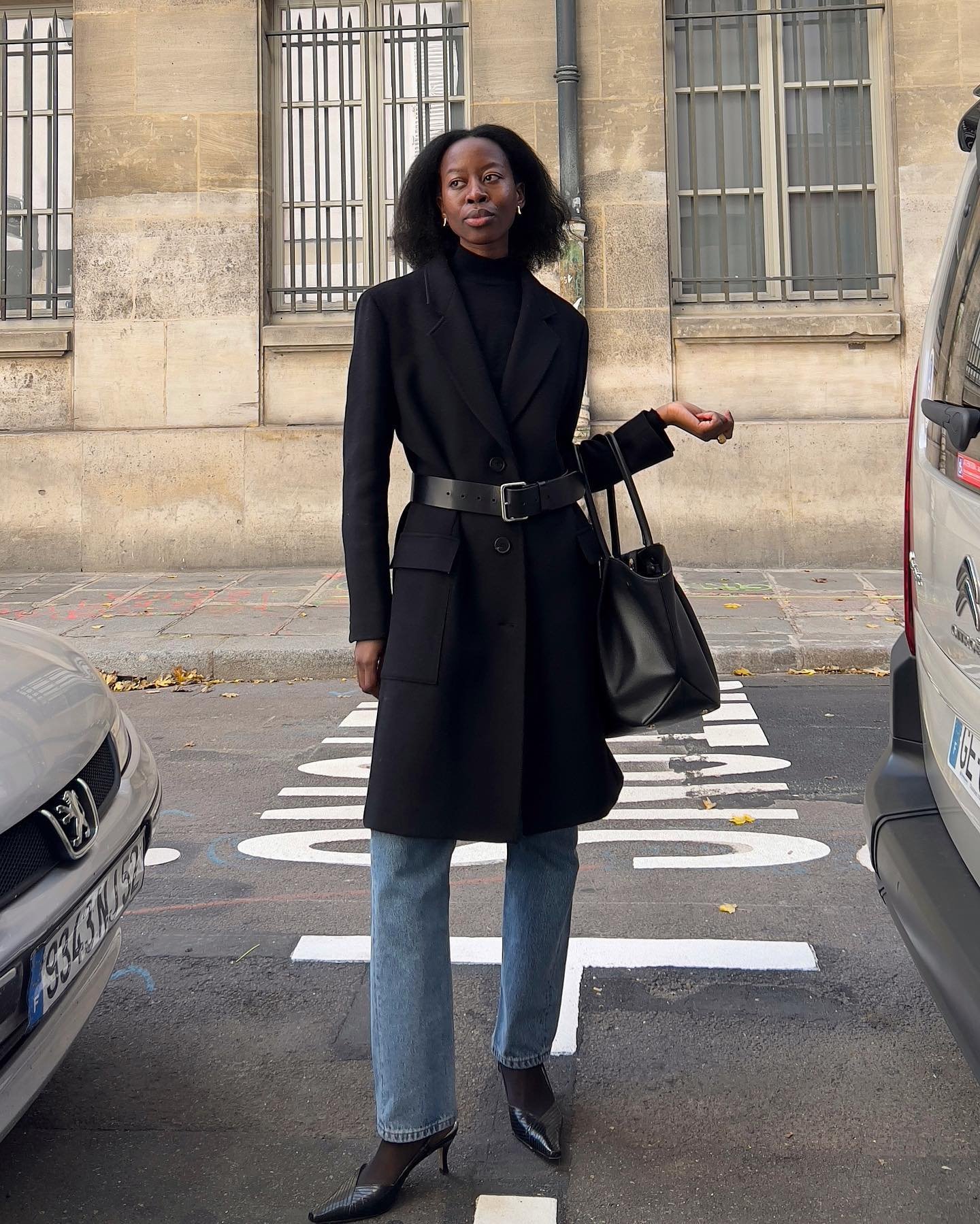 Mulher vestindo casaco preto com cinto largo sobre suéter preto, calça jeans reta e sapatos de salto agulha. Carrega uma bolsa preta grande. O look combina estilos elegantes e casuais, indicando uma tendência minimalista e sofisticada.