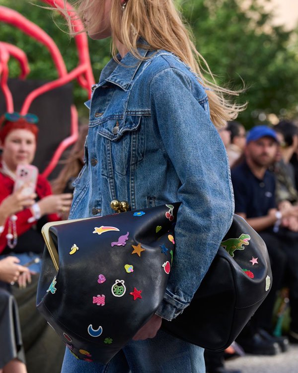 Modelo vestindo jaqueta jeans casual, carregando bolsa preta adornada com patches coloridos e divertidos. O look combina o denim clássico com acessórios ousados e modernos, refletindo tendência jovem e descolada.