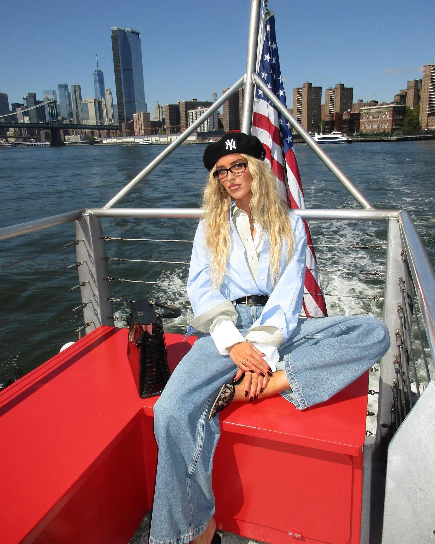 Mulher usando jeans largo, camisa azul clara com mangas dobradas, cinto preto, boina estilosa preta com logo dos Yankees e sapatos de salto fino com estampa de renda. Estilo casual-chic, destacando tendências urbanas e esportivas. cenário de cidade ao fundo.