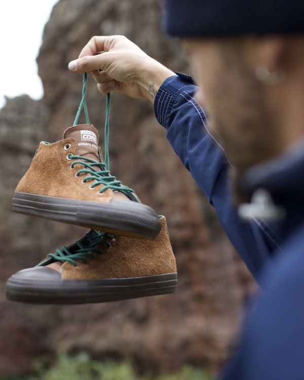 Pessoa segurando tênis marrom de camurça com cadarços verdes e solado robusto. Combinação ideal para estilo casual e aventureiro. Tendência de moda outono-inverno com foco em materiais naturais e duráveis.