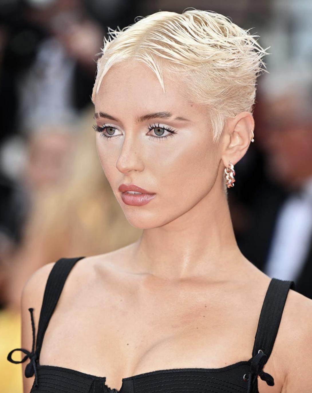 Mulher com cabelo platinado curto, estilo pixie, veste um top preto de alças largas. Maquiagem suave com destaque para os cílios e lábios, acompanhada de um brinco discreto. Estilo moderno e sofisticado, refletindo tendências minimalistas e ousadas na moda.  corte de cabelo curto pixie