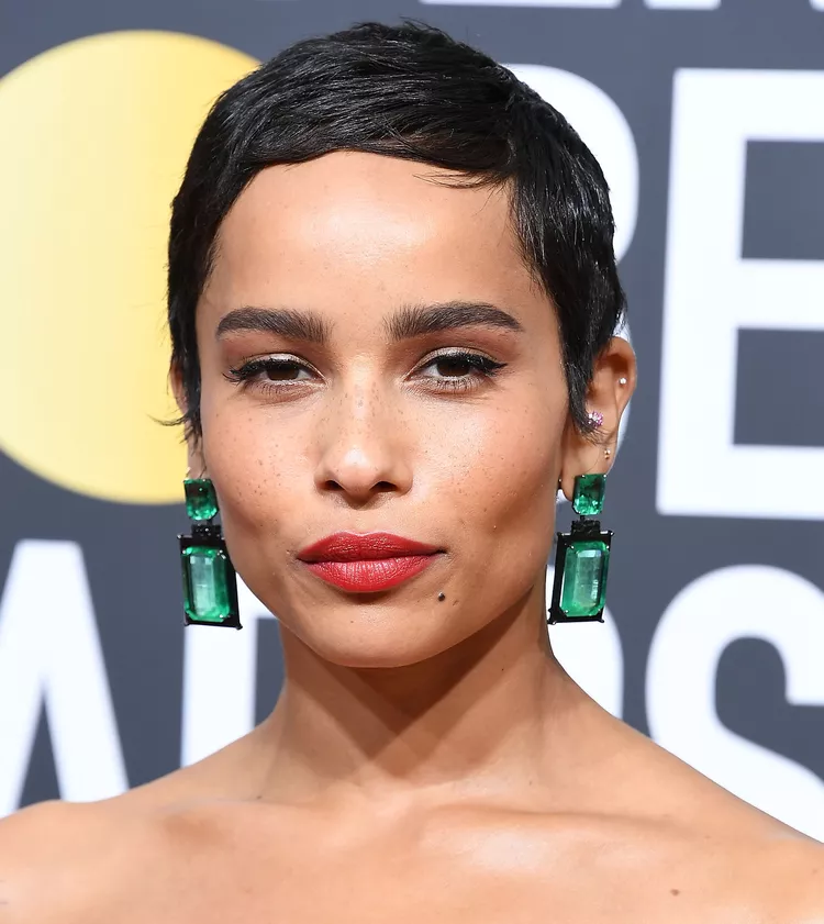 Close-up de uma mulher com cabelo curto e preto, usando brincos grandes com pedras verdes retangulares. Ela está maquiada com batom vermelho vibrante e delineado de olhos clássico. O visual combina elementos modernos e sofisticados, destacando acessórios chamativos.  corte de cabelo curto pixie