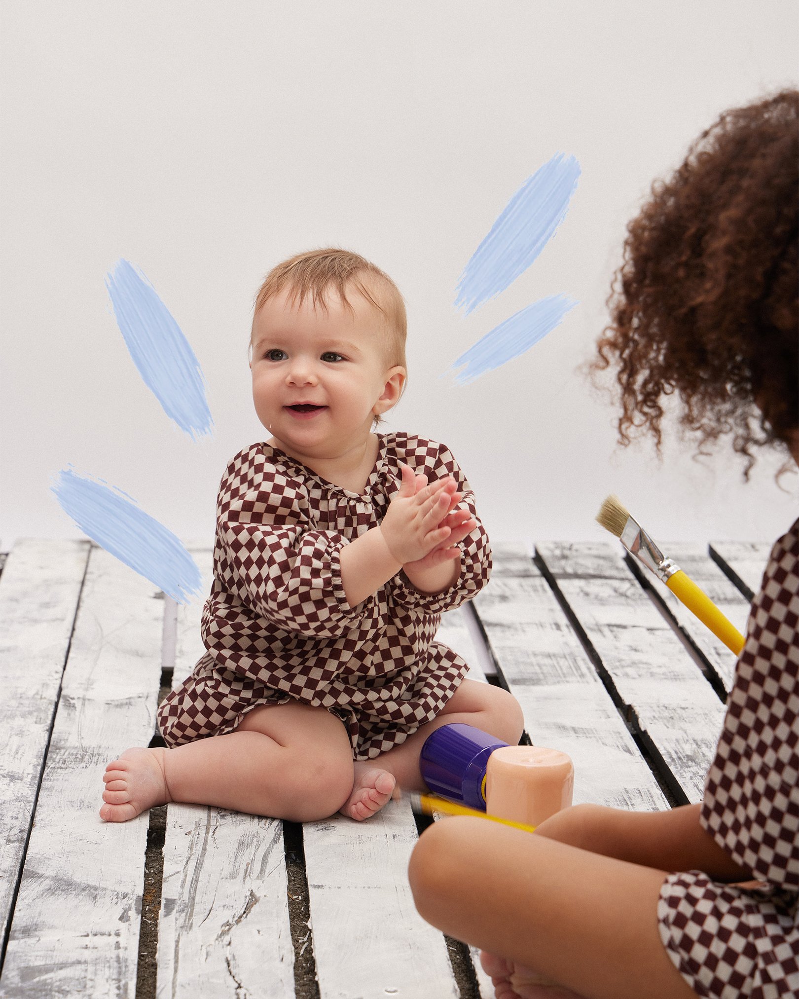 Imagem de bebê sorridente vestindo um macacão xadrez marrom e branco. Ao seu lado, uma criança, também com roupa xadrez, segura pincéis de pintura. Moda infantil com foco em estampas geométricas e cores neutras. Ambiente descontraído e criativo.