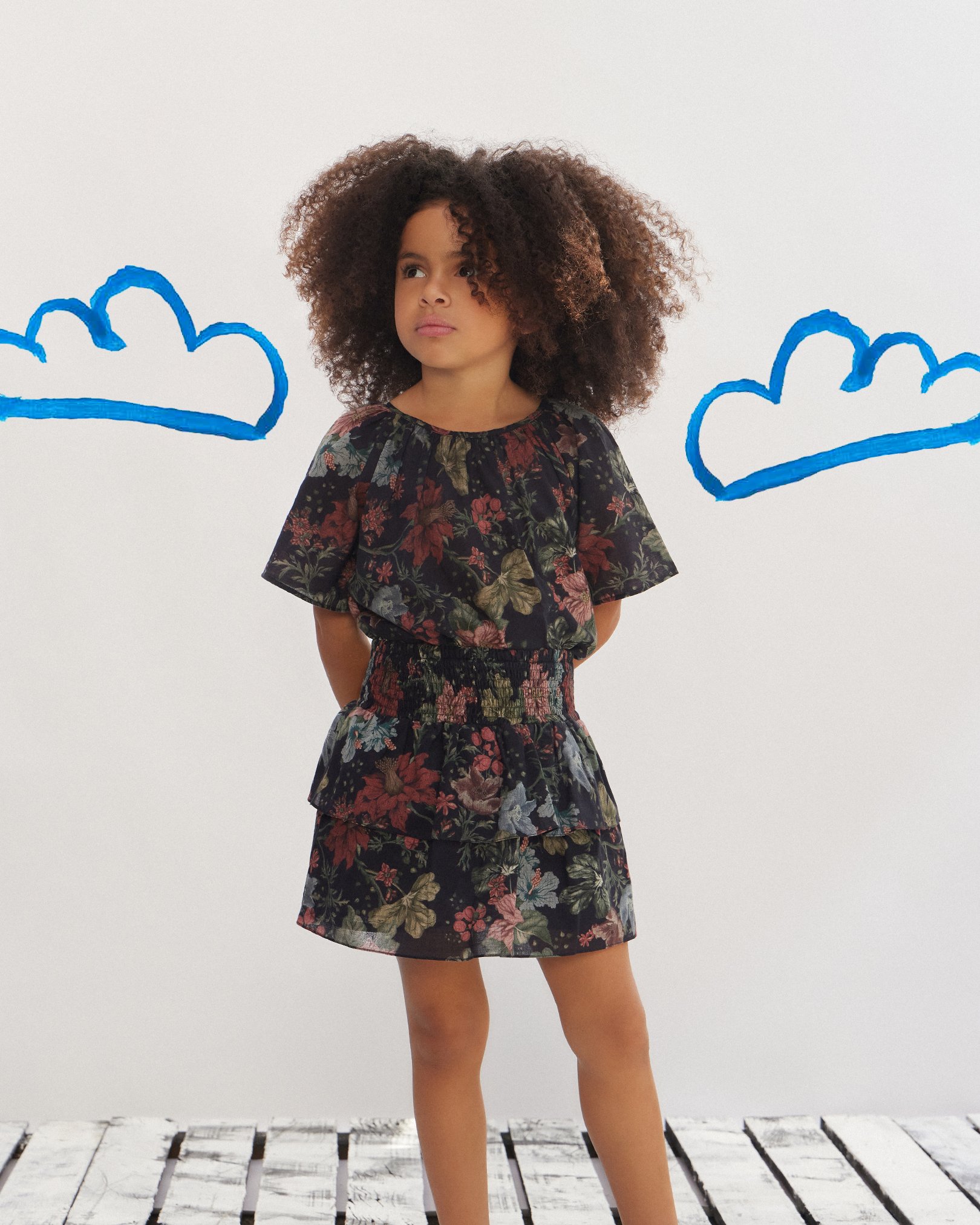 Menina usando vestido floral estampado em fundo escuro, com babados na saia e mangas curtas. Moda infantil atual com influência boho e detalhes românticos, ideal para diversas ocasiões. Estilo descontraído e elegante, destacando-se pelo conforto e charme.