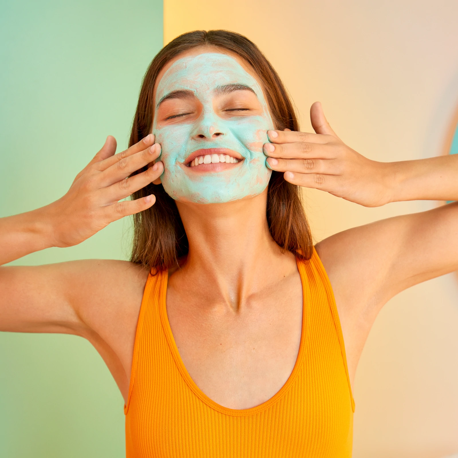 Mulher aplicando máscara facial azul, de kit de skincare para pele oleosa, enquanto veste uma regata laranja vibrante, tendência de moda casual e confortável. A imagem destaca skincare e bem-estar, com cores vivas e atmosfera descontraída.