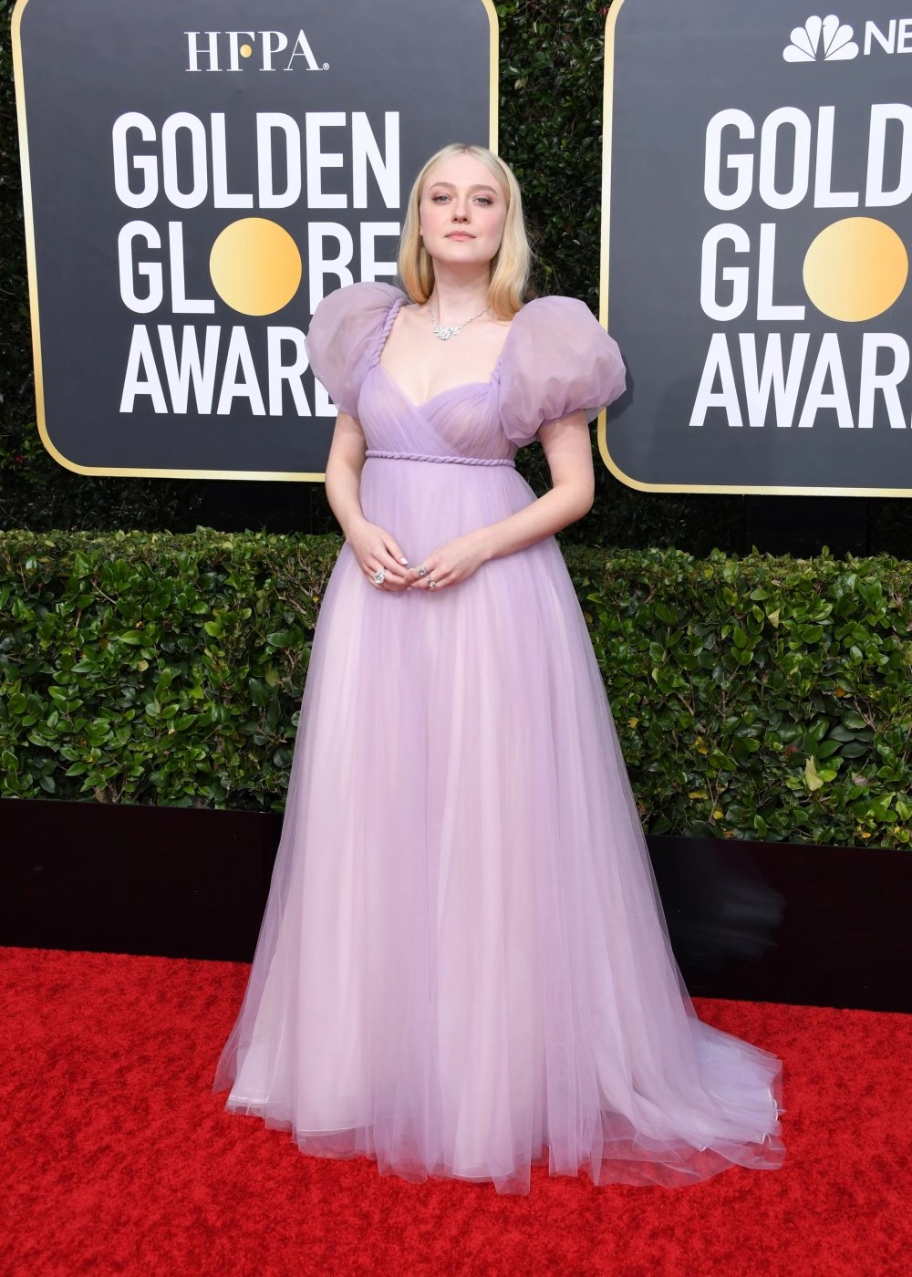 Dakota Fanning usando um vestido longo lilás, com mangas bufantes e detalhe franzido na cintura, seguindo uma tendência romântica e delicada. Look sofisticado e elegante, perfeito para eventos formais, como premiações.