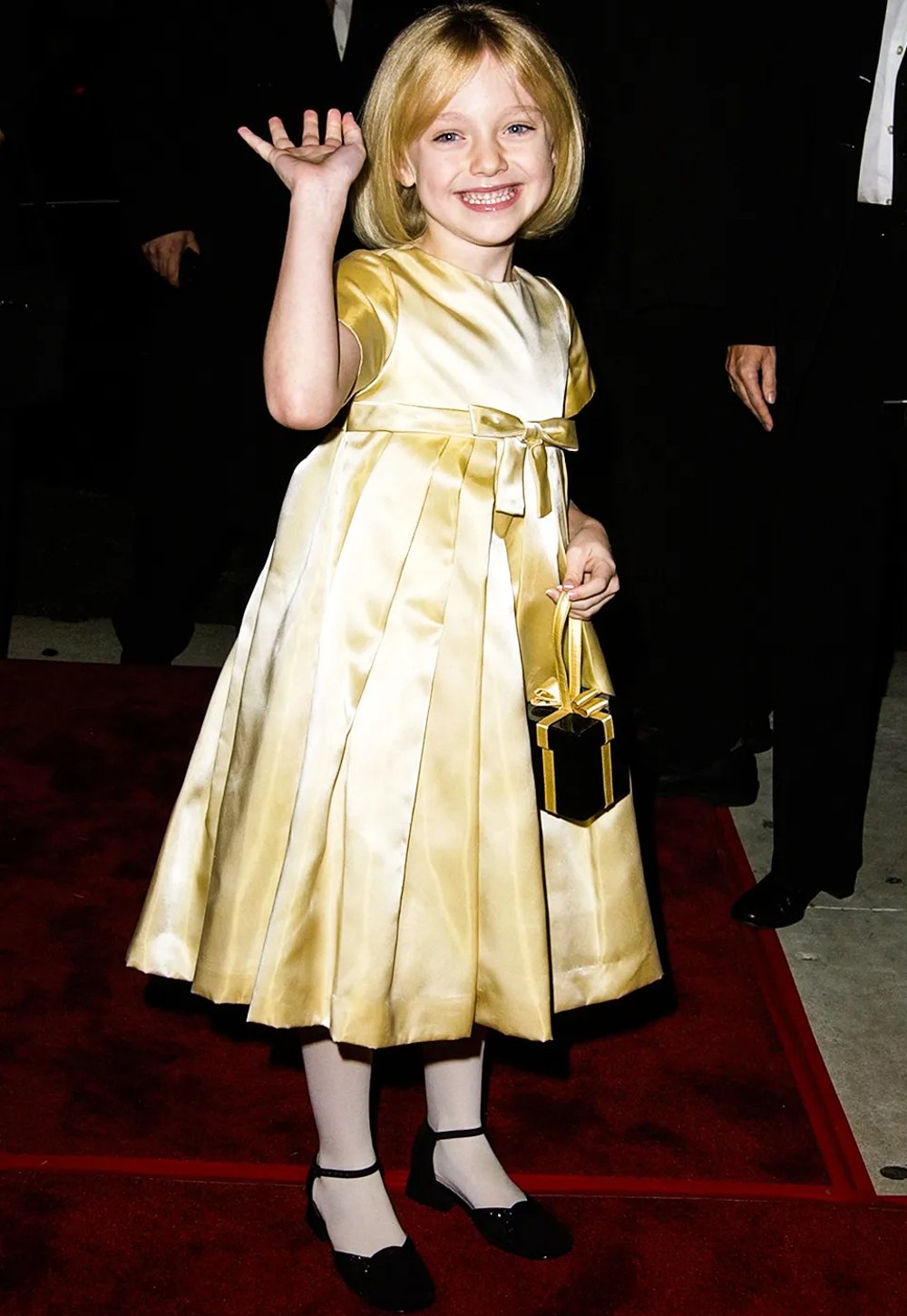 Dakota Fanning usando vestido dourado de cetim com laço na cintura, meias brancas e sapatos pretos, completando o look com bolsa de mão preta com detalhes dourados. O estilo remete a um visual clássico e elegante, ideal para eventos formais.