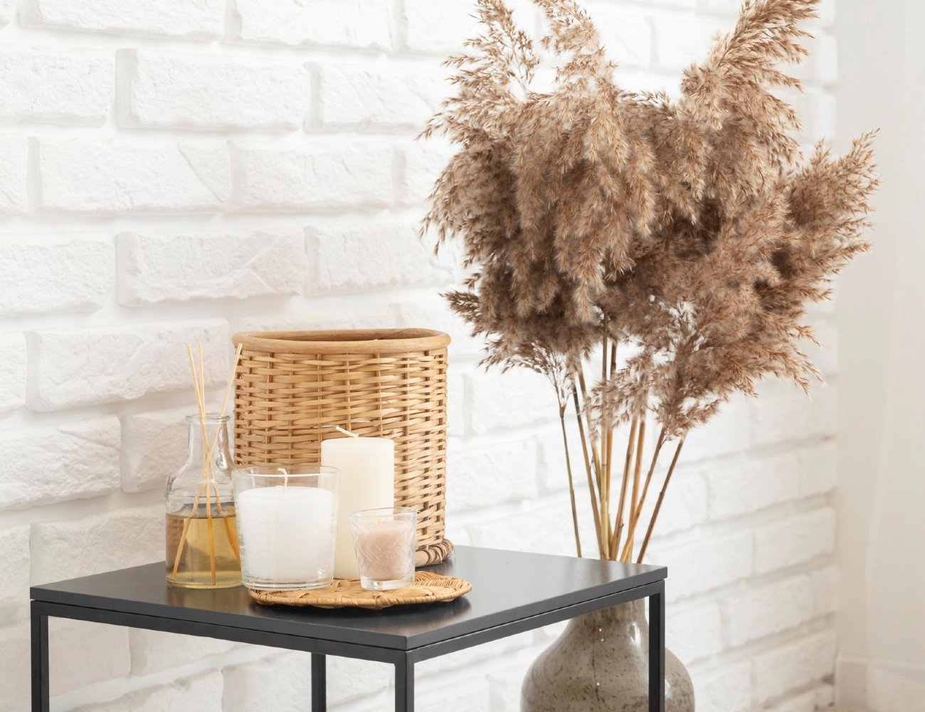 Imagem mostrando uma mesa decorativa com itens minimalistas: vaso de vidro, velas brancas, cesta de palha e arranjo seco de capim-dos-pampas. Tendências de design de interiores focadas em decoração escandinava e boho chic, com tons neutros e materiais naturais.