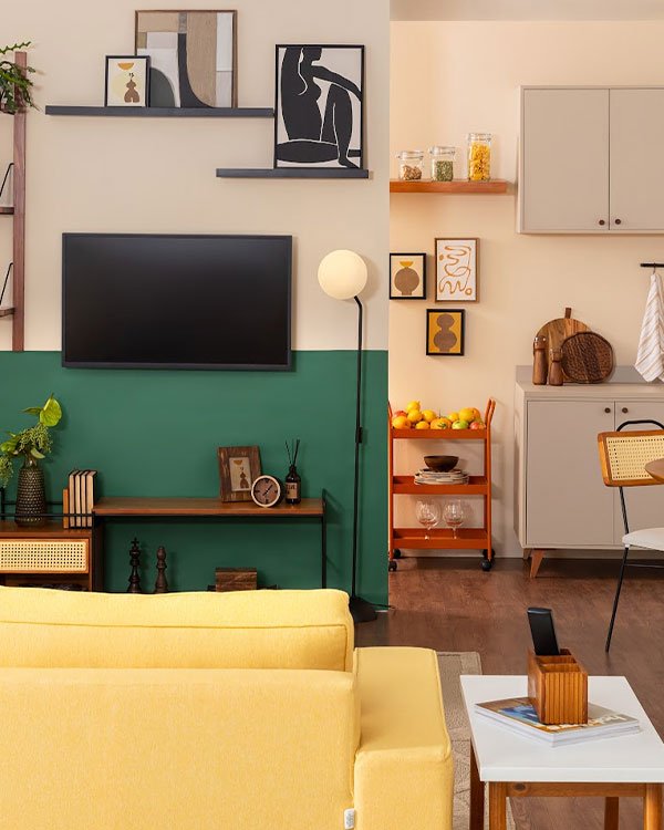 Sala de estar moderna e estilosa com sofá amarelo, parede verde e creme dividida por meia pintura, quadros minimalistas e decoração contemporânea. Estante aberta com objetos decorativos, carrinho de bar laranja e móveis de madeira neutros.