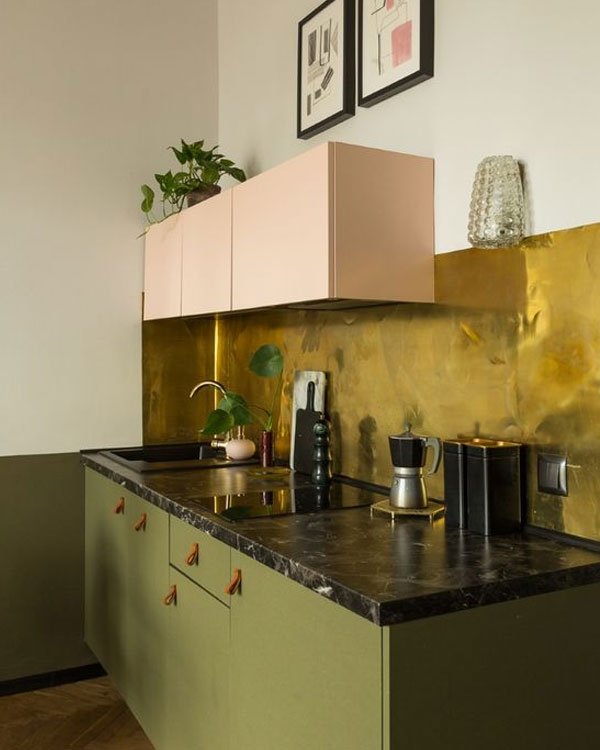 Cozinha moderna com armários inferiores verde oliva e superiores em rosa claro. Bancada de mármore preto e parede dourada destacam um design sofisticado e contemporâneo.