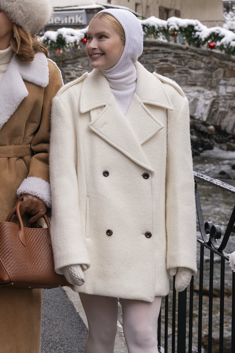 Imagem de duas mulheres em ambiente nevado, uma delas veste um sobretudo de lã branco com botões escuros, combinando com um gorro de malha branco ajustado à cabeça. Quarta temporada de Emily em Paris.