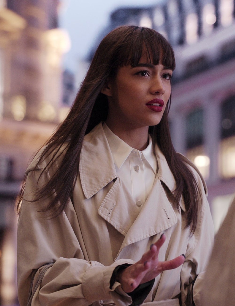 Mulher usando um trench coat bege sobre uma camisa branca. O cabelo é longo e liso, com franja reta. Look clássico e atemporal, ideal para climas frescos. Quarta temporada de Emily em Paris.