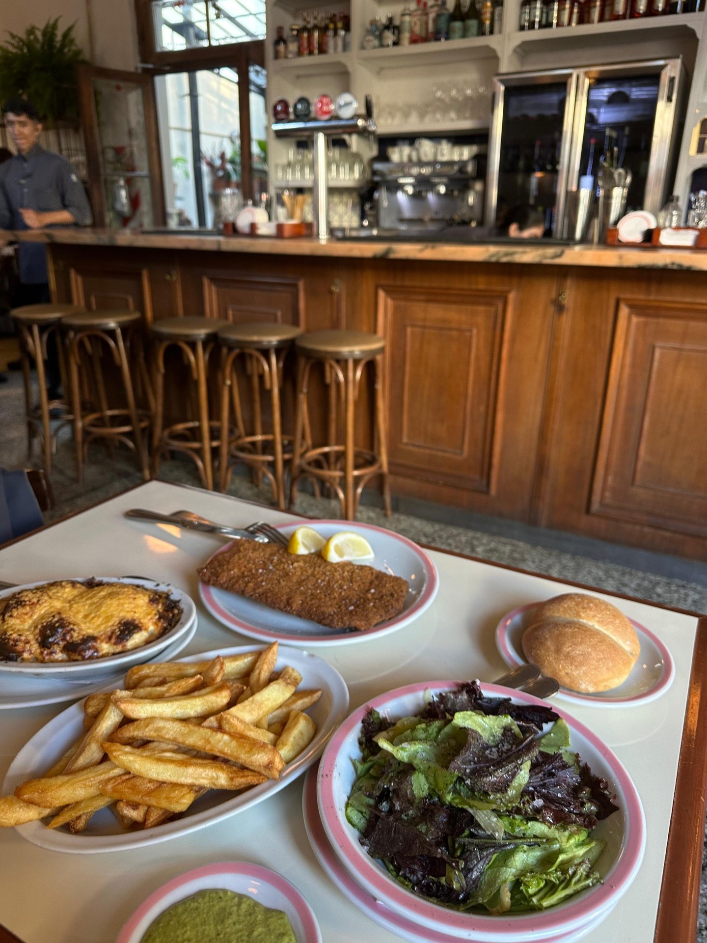 Imagem de restaurante aconchegante com balcão de madeira, banquetas de vime e decoração rústica. Na mesa, refeições incluem salada, batata frita, carne empanada e pães. Ambiente remete a bistrô europeu, com moda casual e charme clássico.