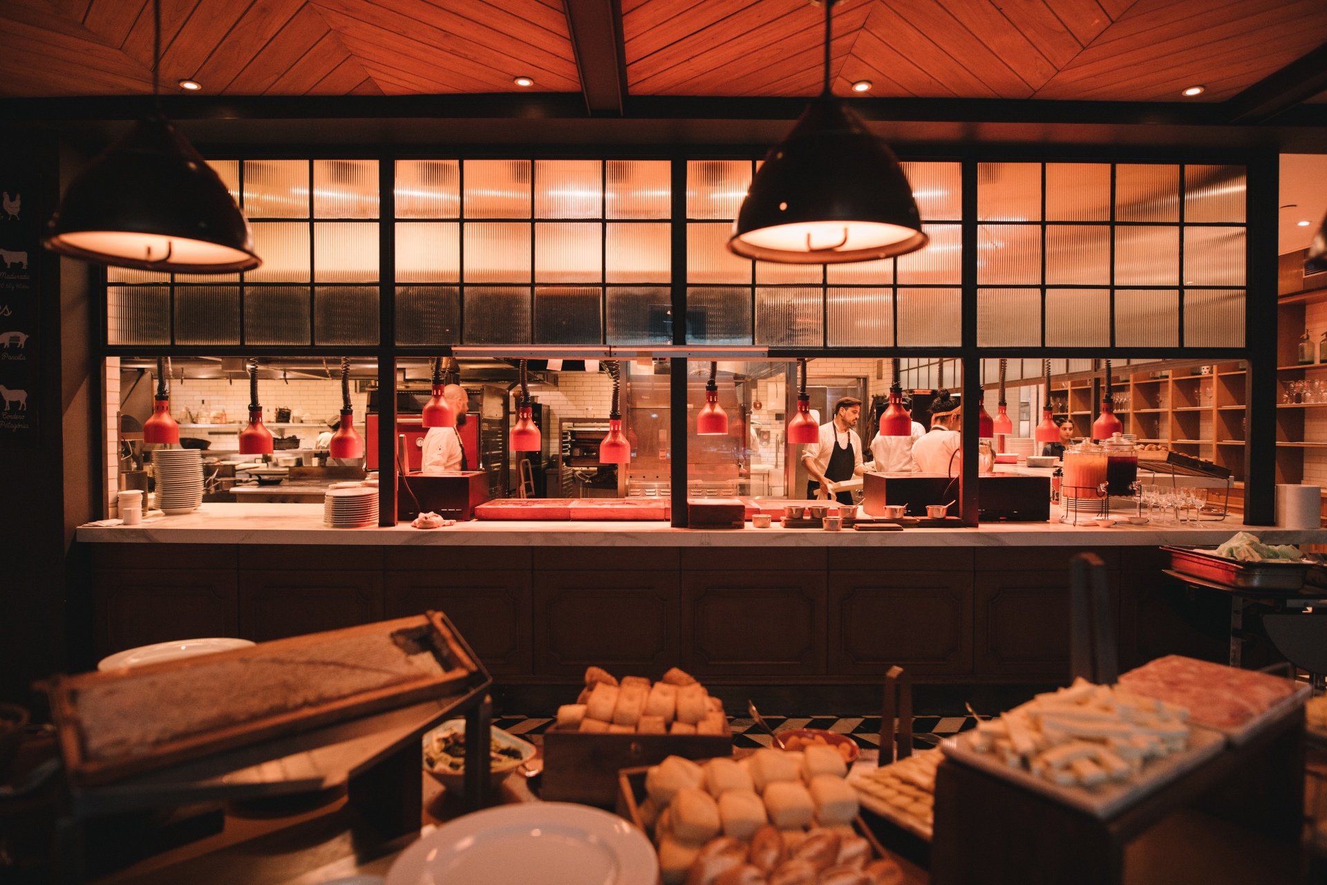 Restaurante com cozinha aberta. Chefs e cozinheiros vestem uniformes tradicionais: dólmãs brancas, aventais pretos e chapéus de cozinha, seguindo uma estética clássica e profissional. Ambiente moderno com luminárias pendentes e design rústico-industrial.