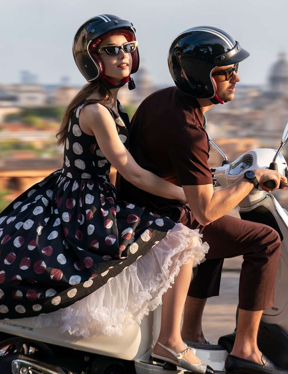 Casal em scooter. Mulher usa vestido vintage de bolinhas, armação interna volumosa, sapatos baixos e óculos de sol estilo retrô. Homem com camisa polo marrom, calça ajustada e sapatos. Quarta temporada de Emily em Paris.