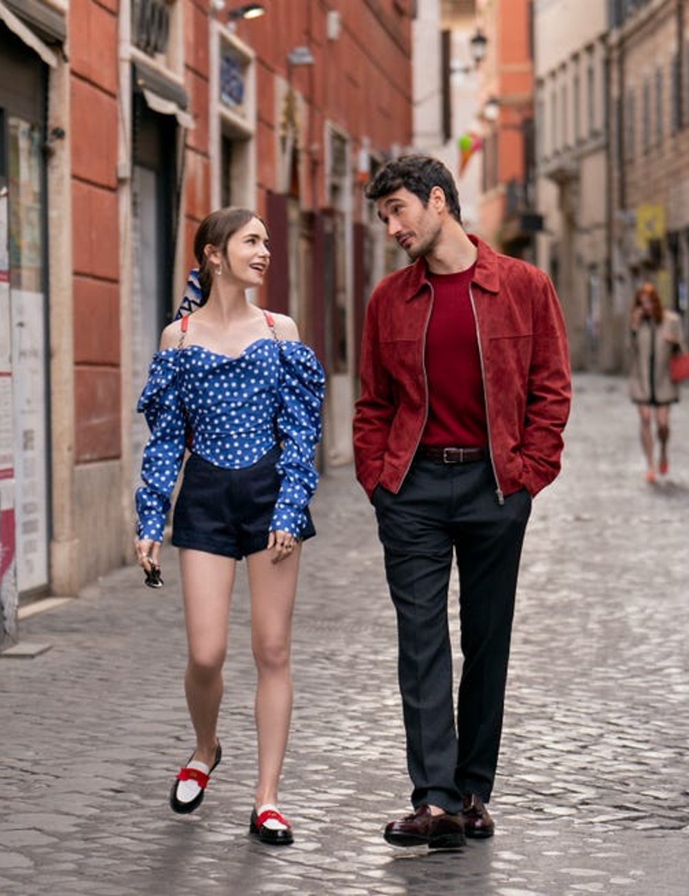 Casal caminhando por rua europeia. Ela usa blusa azul de bolinhas com ombros de fora, shorts pretos e mocassins bicolores. Ele veste jaqueta marrom-avermelhada, suéter vermelho e calça preta. Quarta temporada de Emily em Paris.