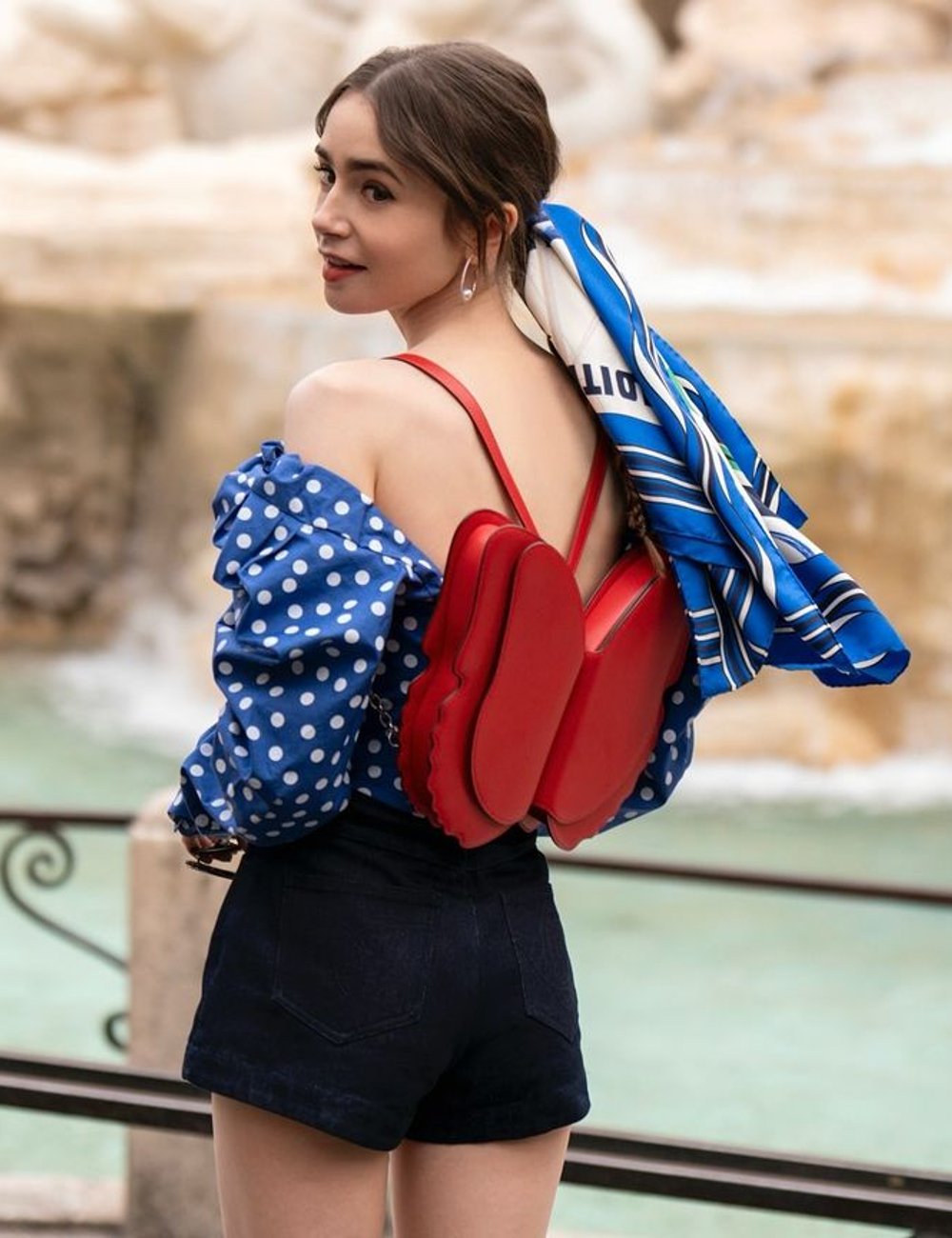 Jovem usa blusa azul de bolinhas brancas com mangas bufantes, short preto de cintura alta e mochila vermelha. Lenço azul amarrado no cabelo complementa o look. Quarta temporada de Emily em Paris.